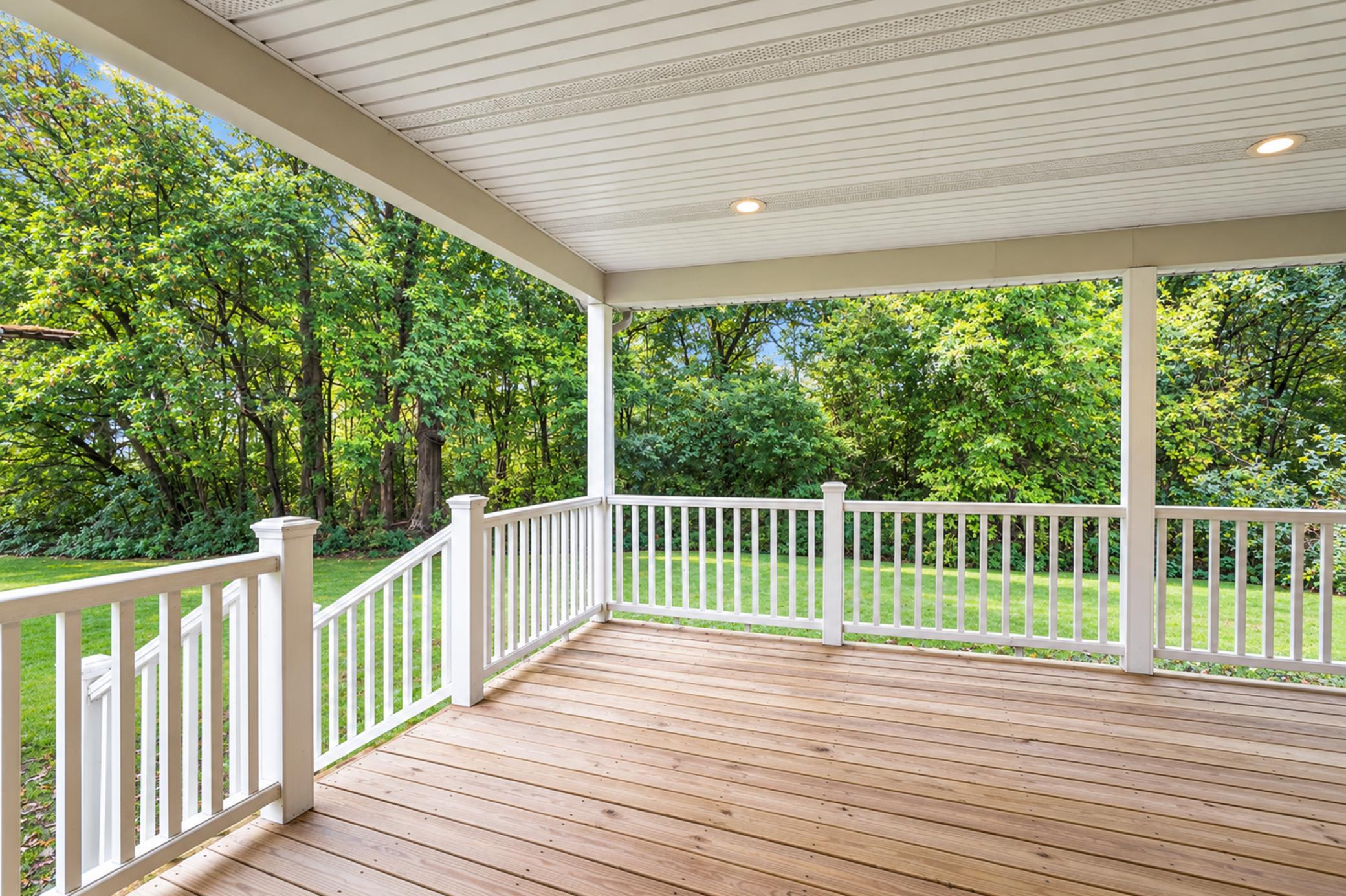 Covered wooden deck with white railings overlooking a green lawn and leafy trees.