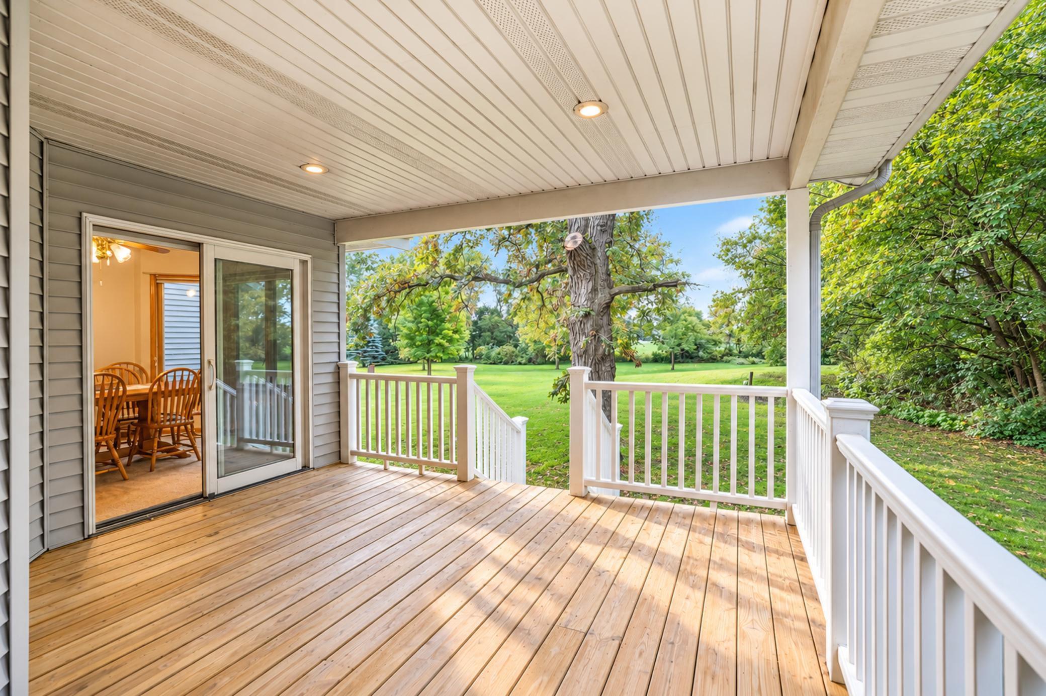 Covered wood deck with white railings overlooking a leafy lawn and mature trees.
