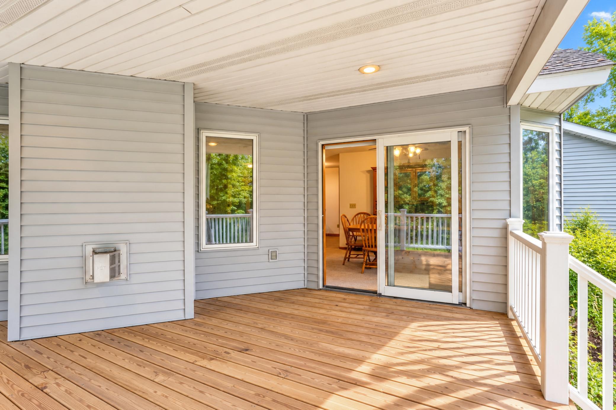 Covered wood deck with white railings, gray siding, and a sliding glass door opening to a dining area.