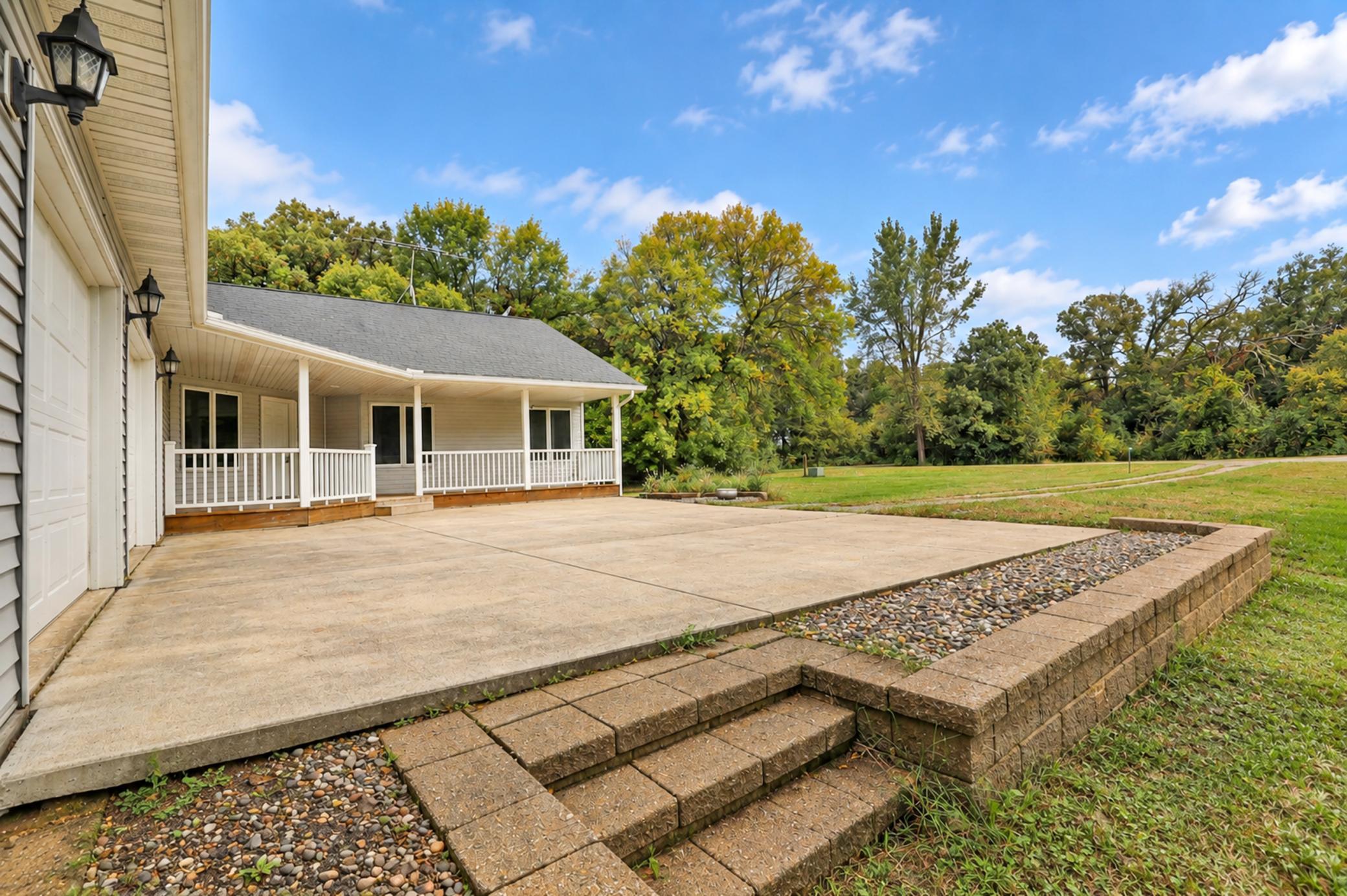 Concrete patio beside a covered porch, with tiered block steps and a tree-lined lawn.