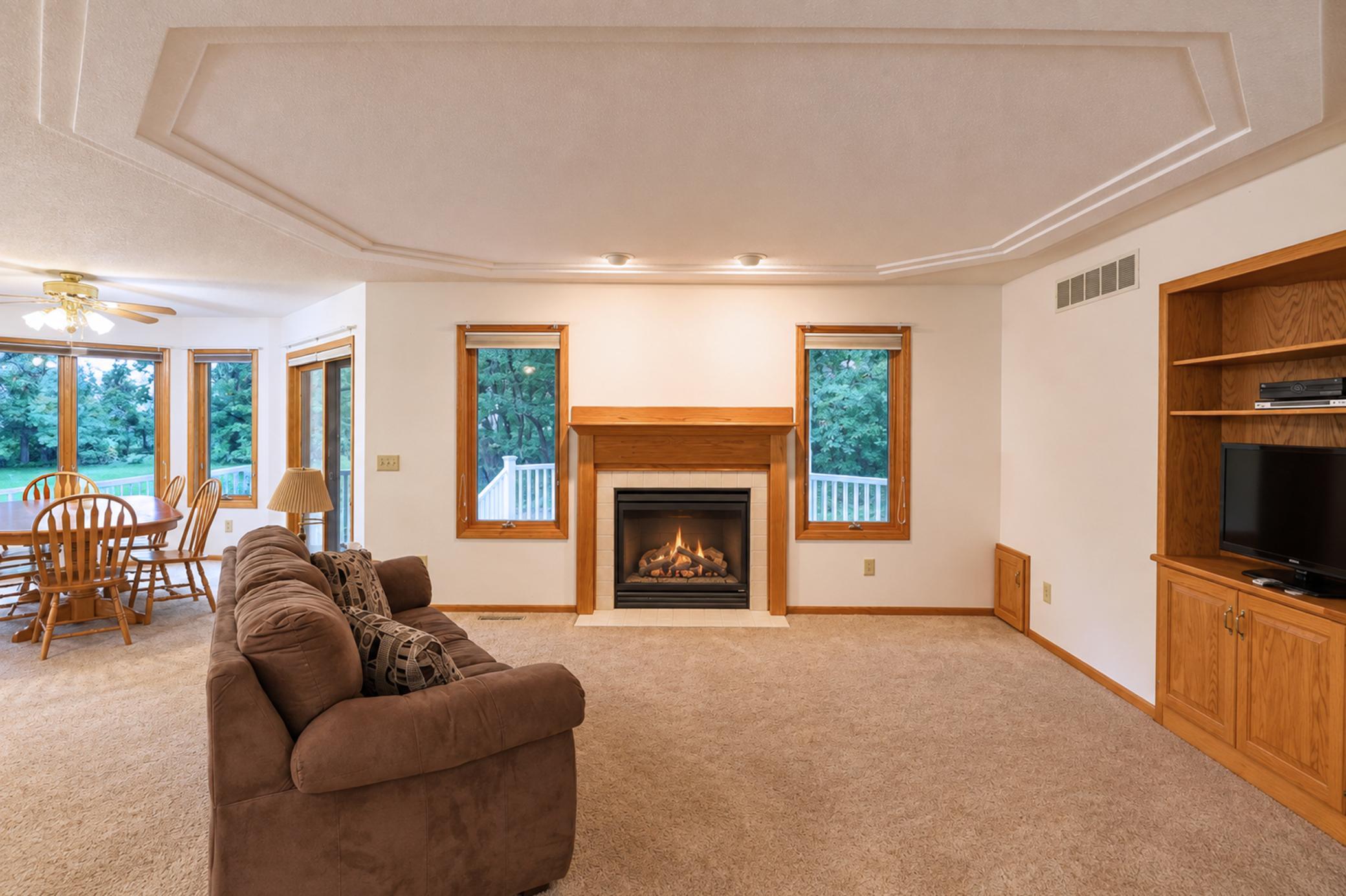 Living room with a wood-trimmed fireplace, brown sofa, and large windows overlooking greenery.