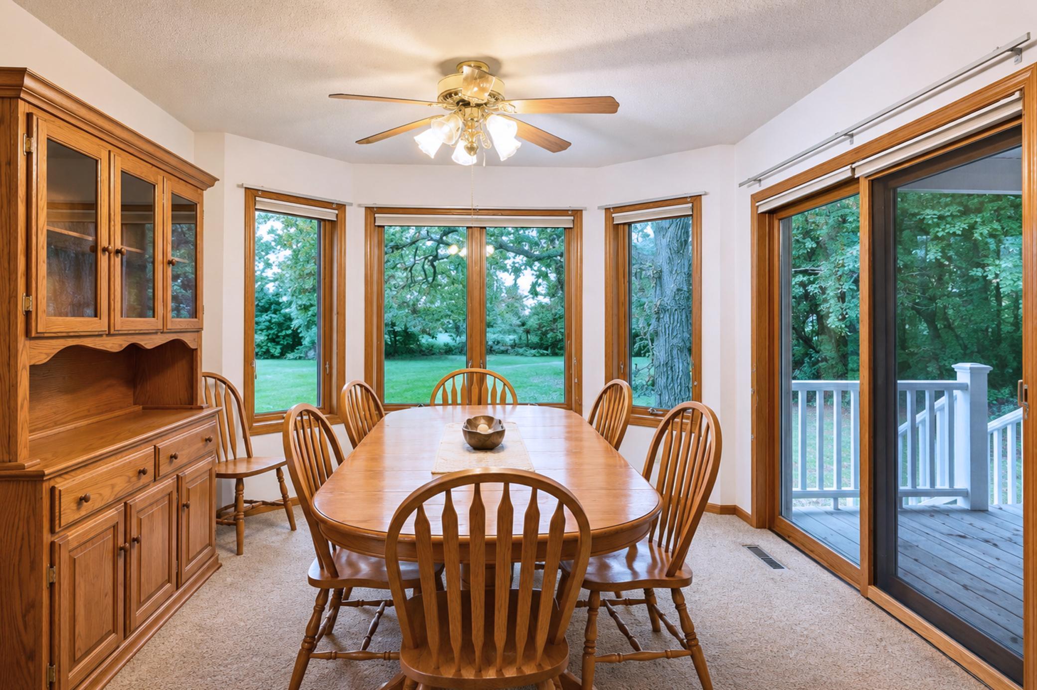 Dining room with a wooden table and built-in hutch, surrounded by windows overlooking a green yard.