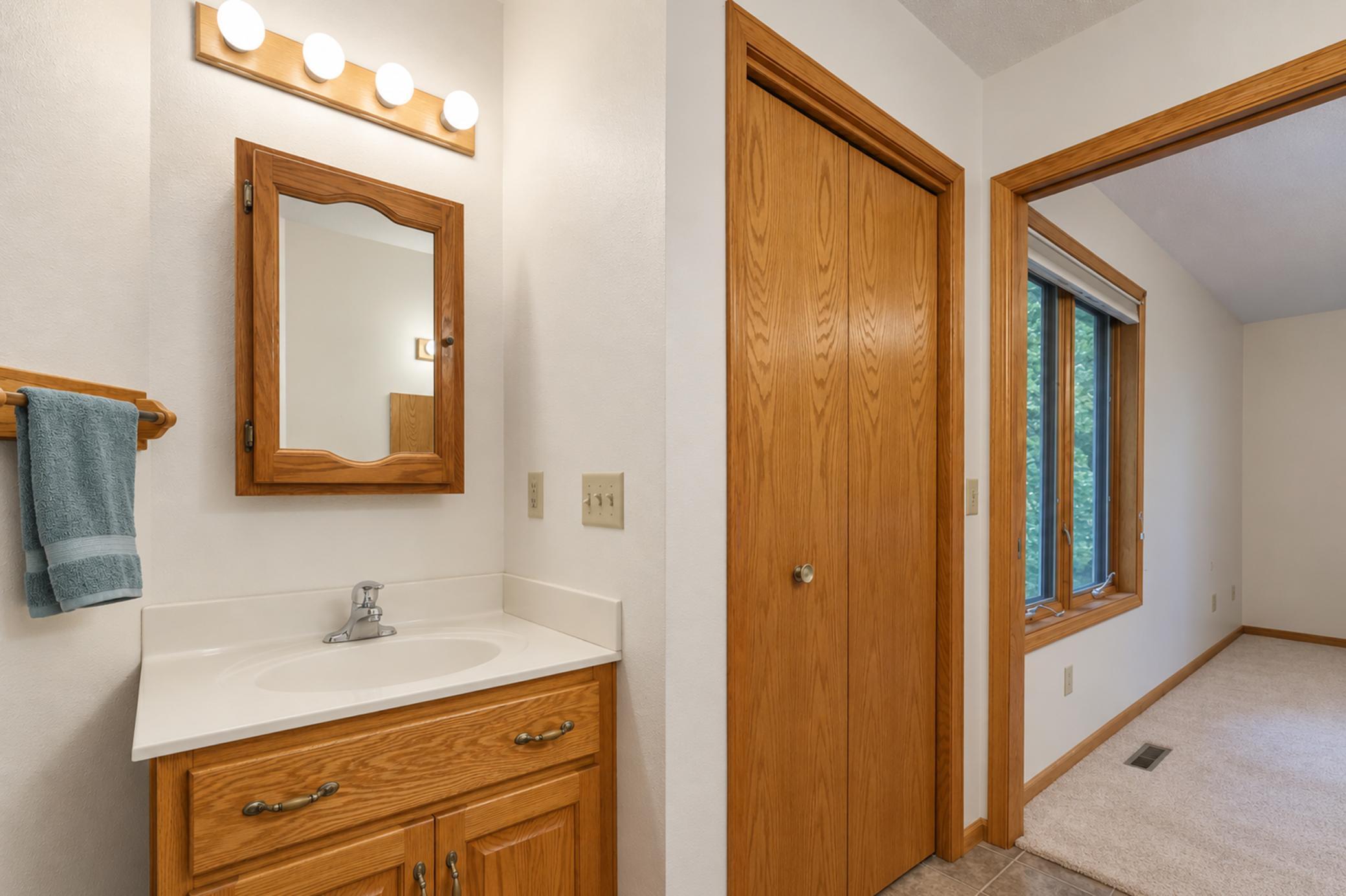 Bathroom vanity with an oak cabinet and mirror beside a doorway to a carpeted room with a large window.