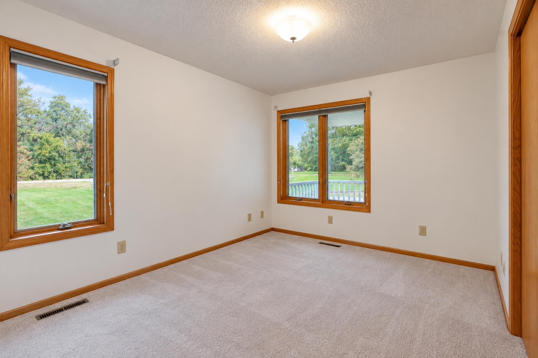 Empty carpeted room with wood-trimmed windows overlooking a green yard.