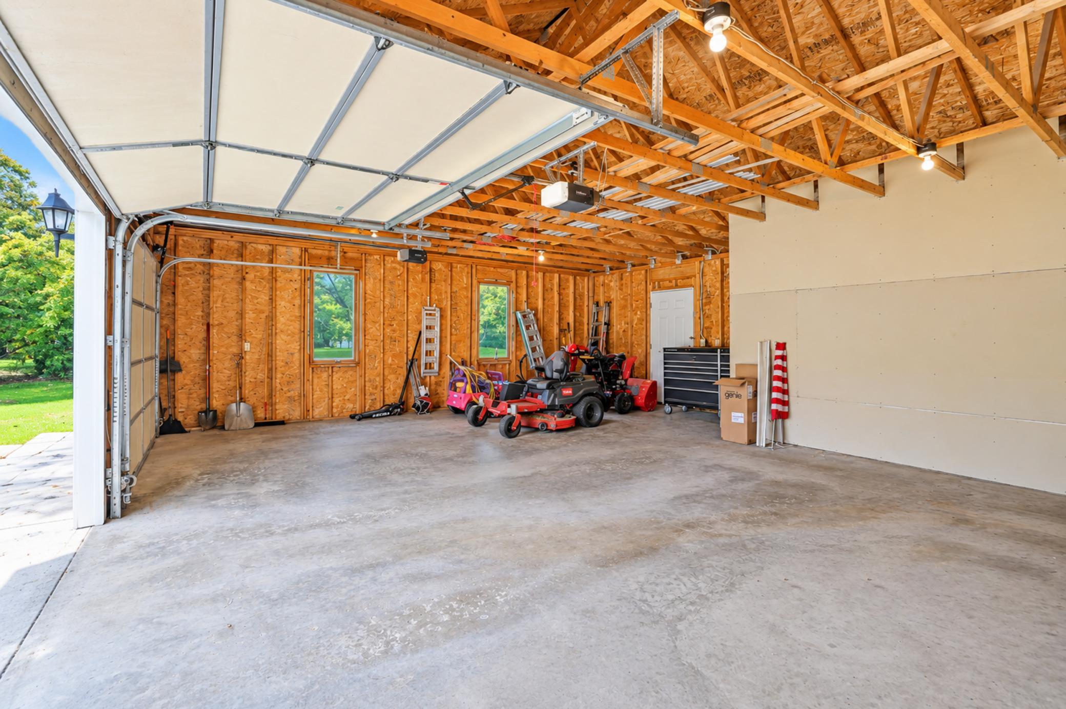 Garage with open wooden trusses, exposed framing, and lawn equipment along the back wall.