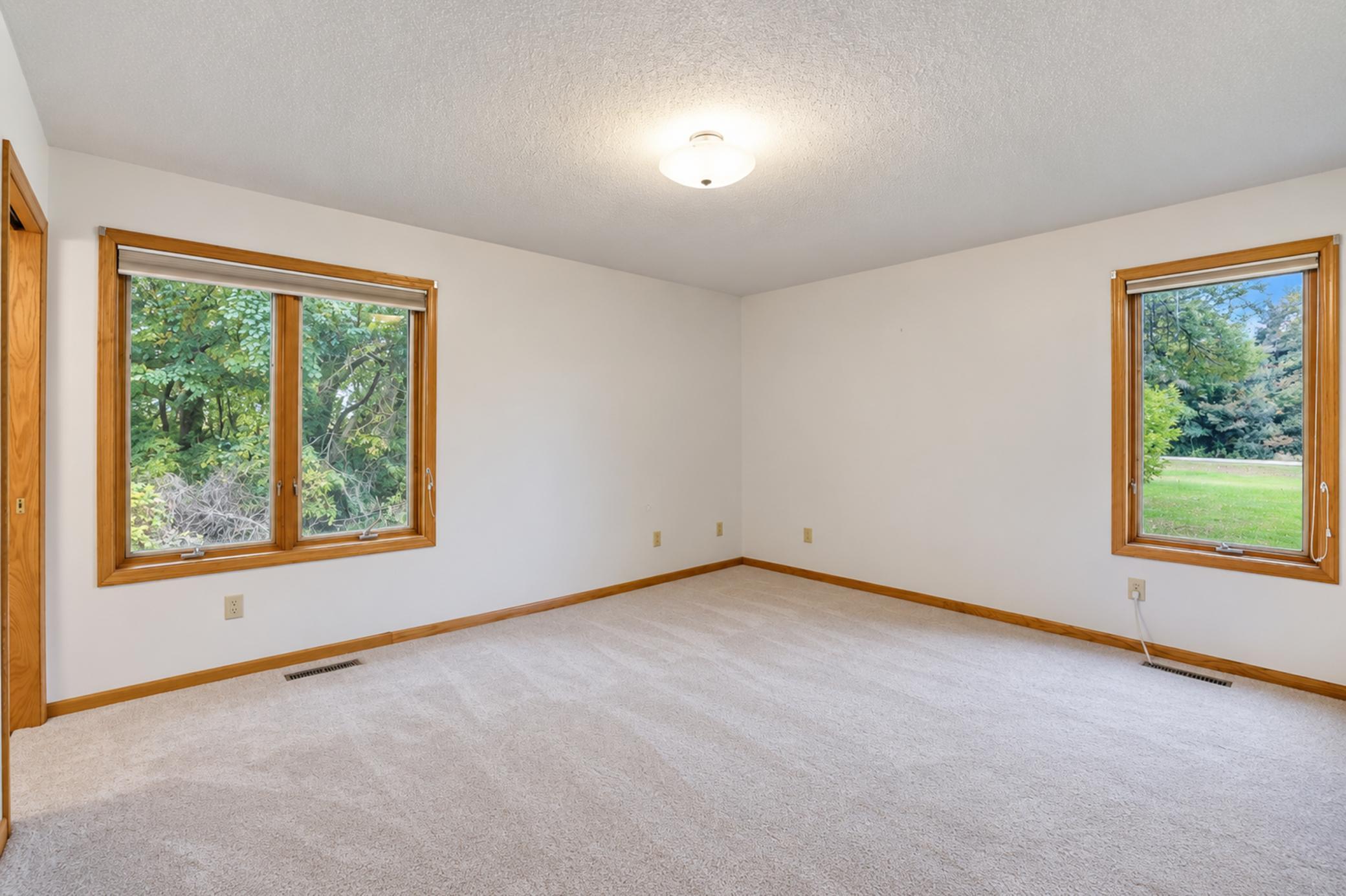 Empty carpeted room with wood-trimmed windows overlooking leafy trees and a lawn.