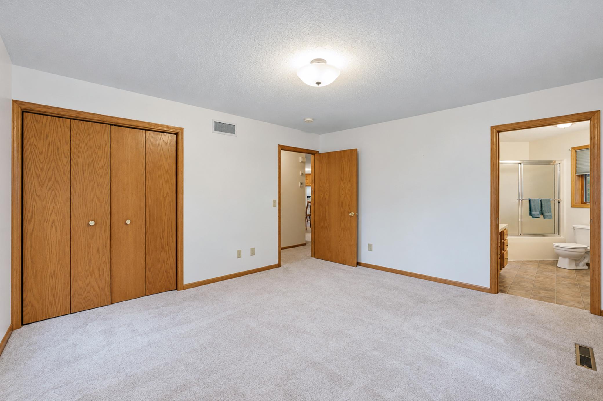 Empty carpeted bedroom with wooden closet doors and an adjoining bathroom.