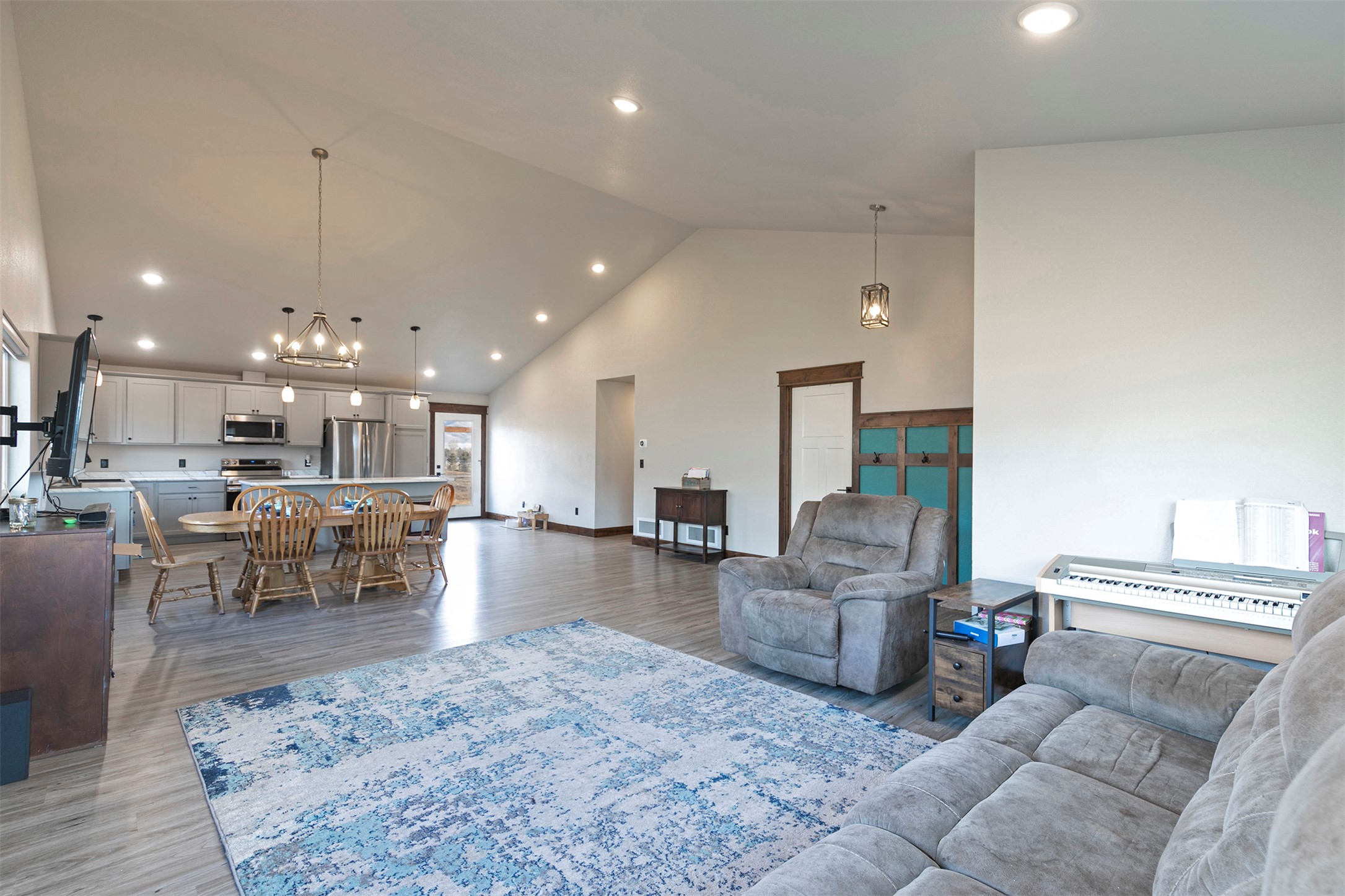 Open-plan living room and kitchen with vaulted ceiling, gray seating, blue rug, and digital piano.