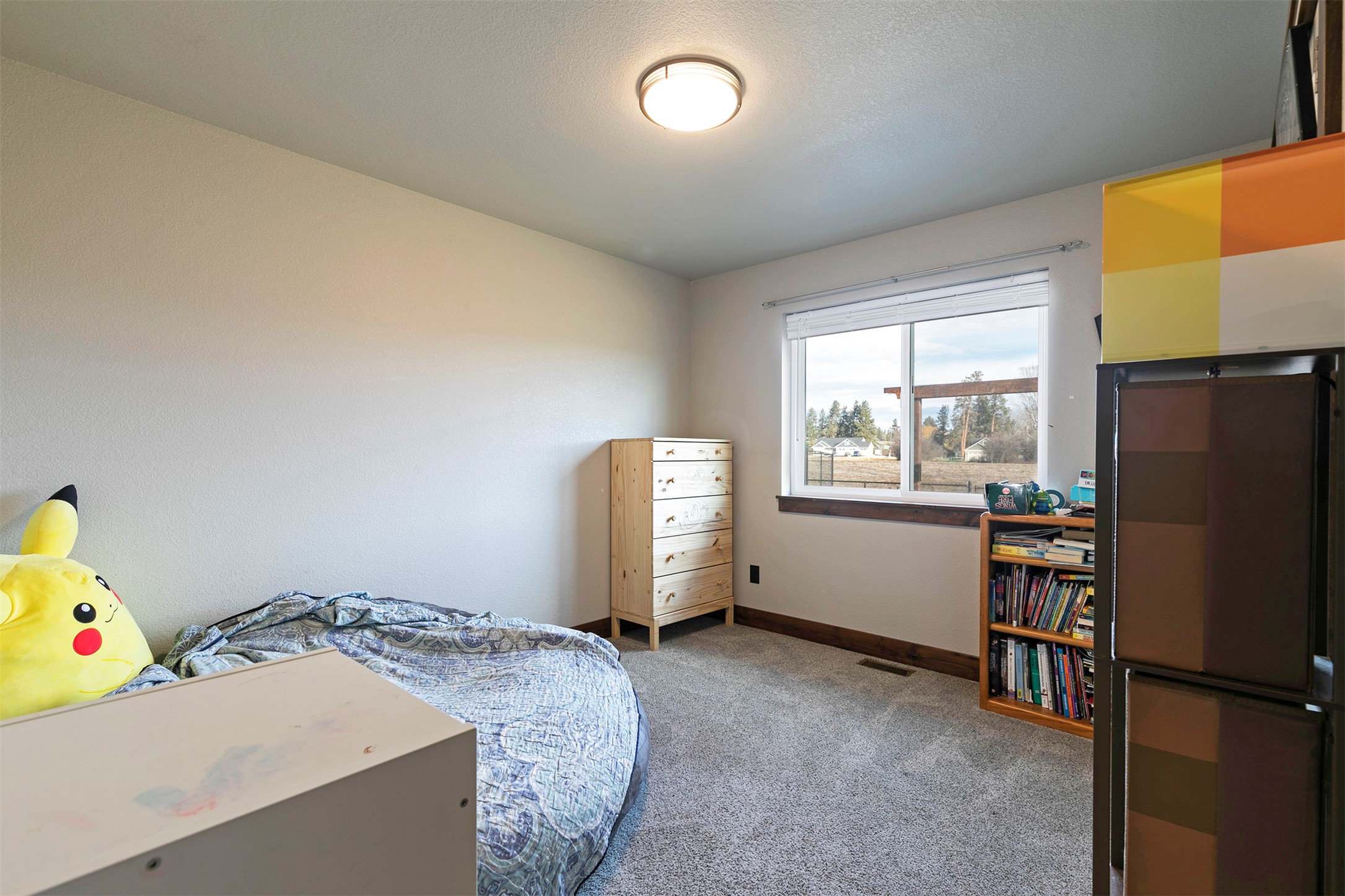Bedroom with a low bed, large Pikachu plush, wooden dresser, and window overlooking a yard.