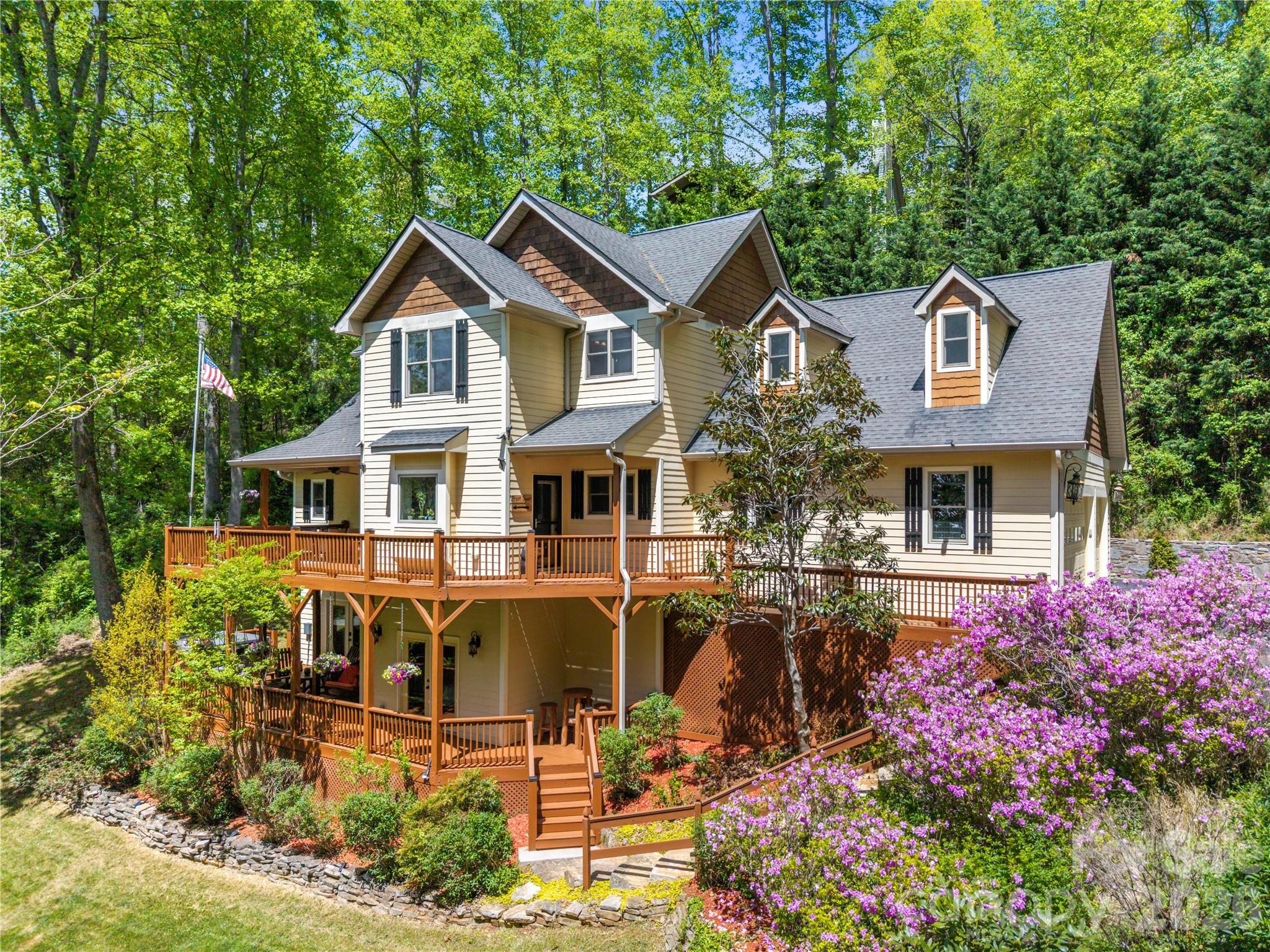 Multi-level cream home with wraparound wooden decks, surrounded by trees and flowering shrubs.