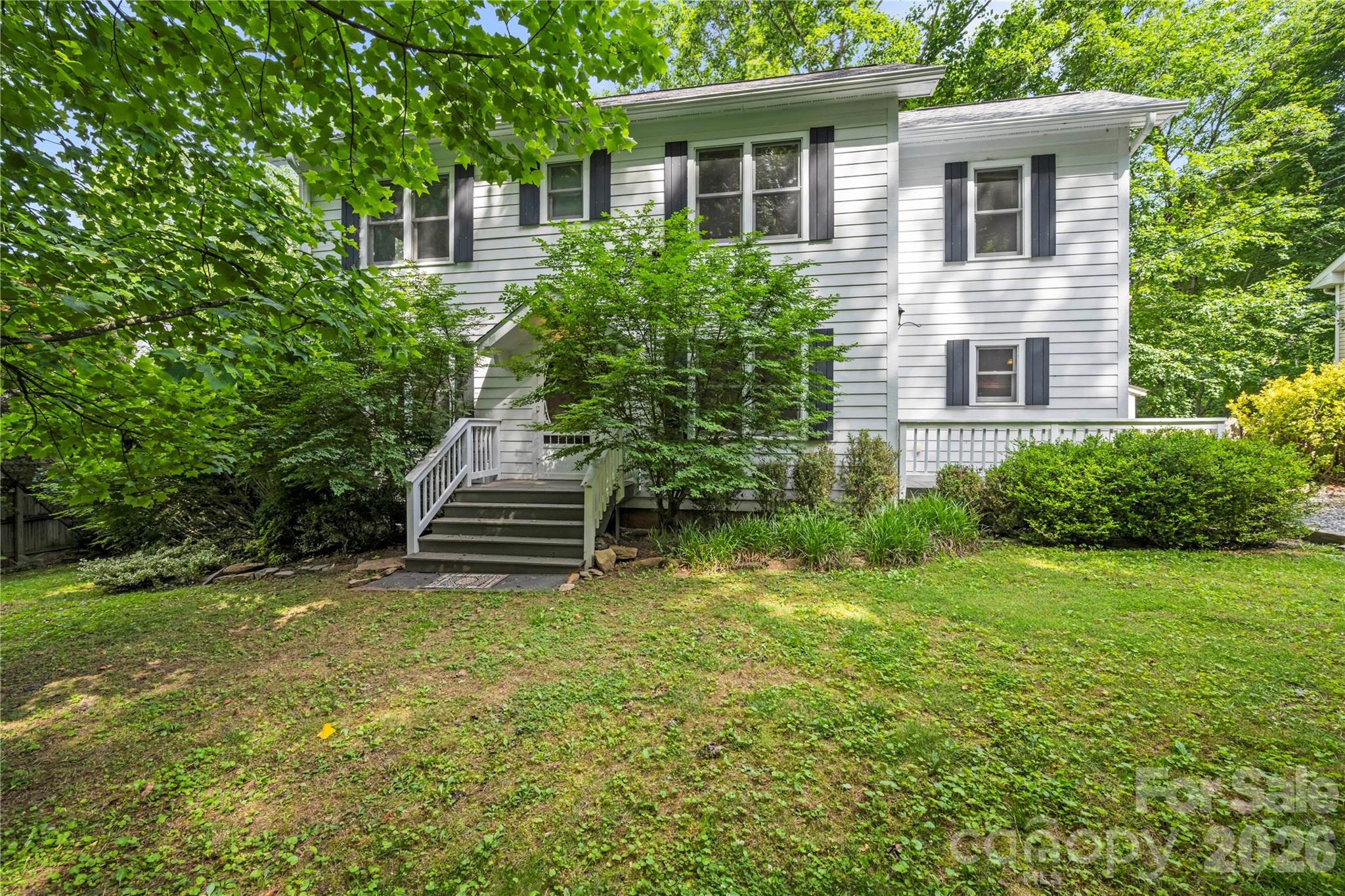 White two-story house with dark shutters and a small staircase, surrounded by leafy trees and shrubs.