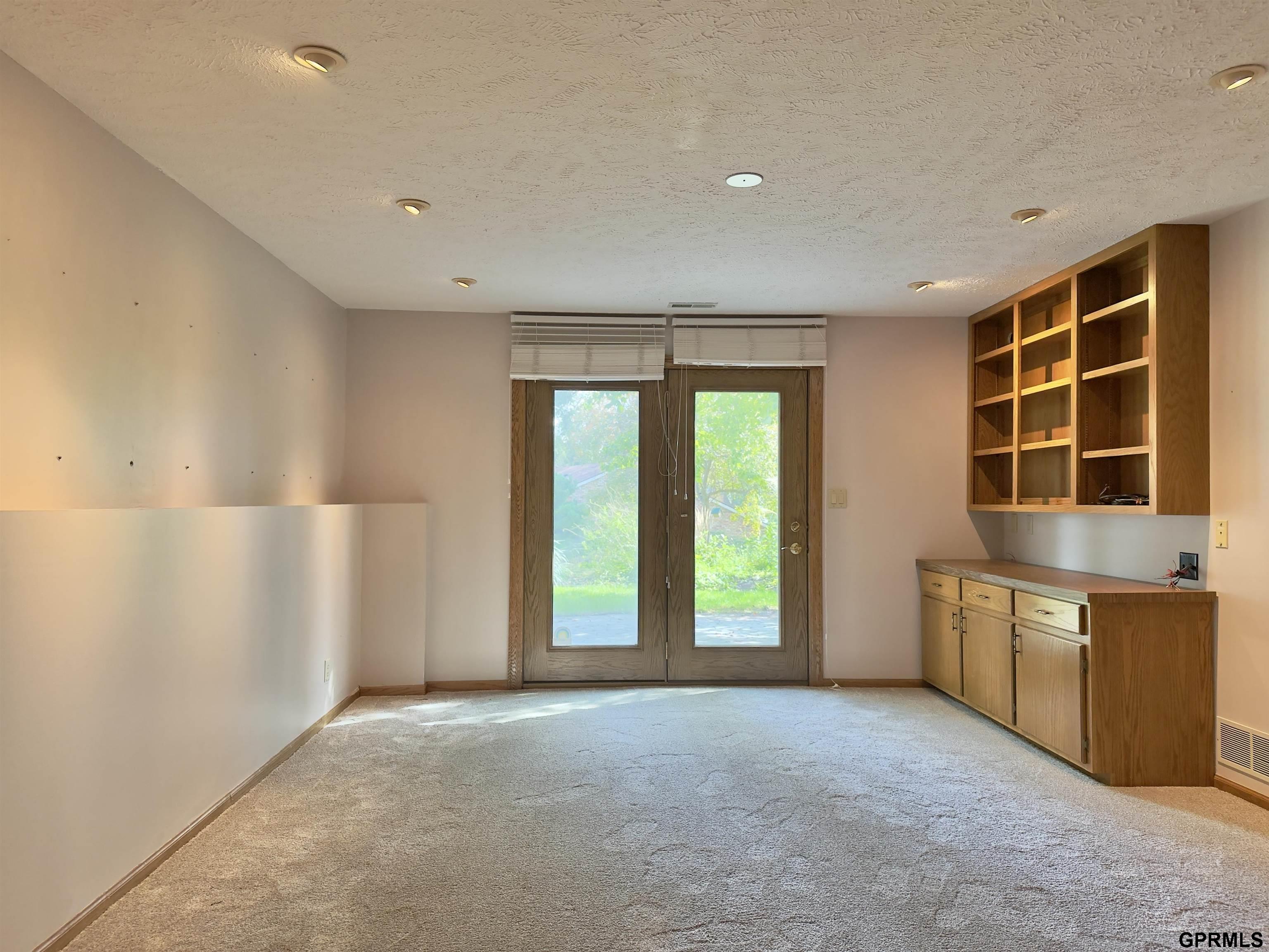 Vacant carpeted room with built-in wooden cabinetry and glass doors opening to a green view.
