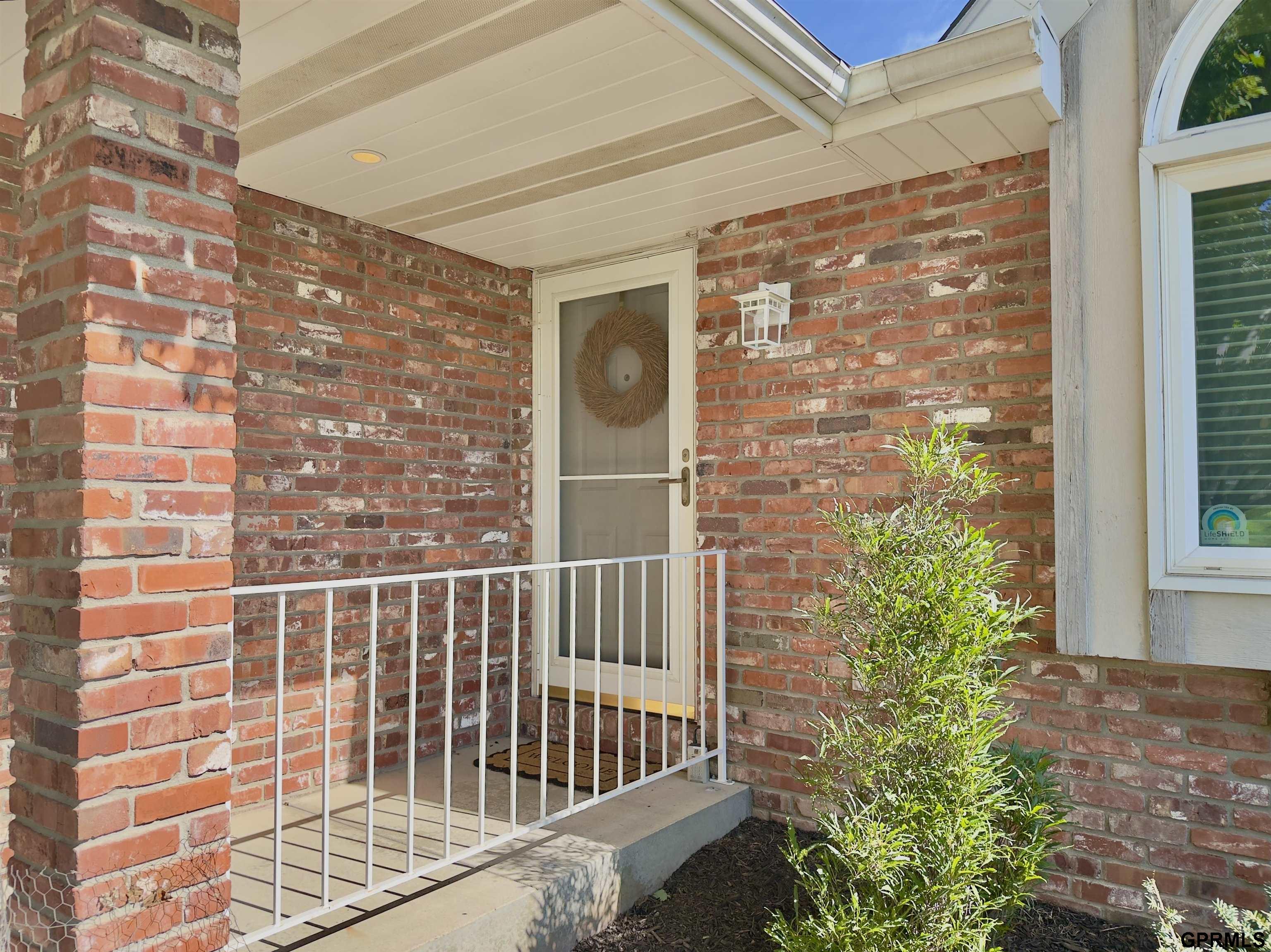 Brick home’s covered front porch with a white railing, wreath on the storm door, and leafy shrub.