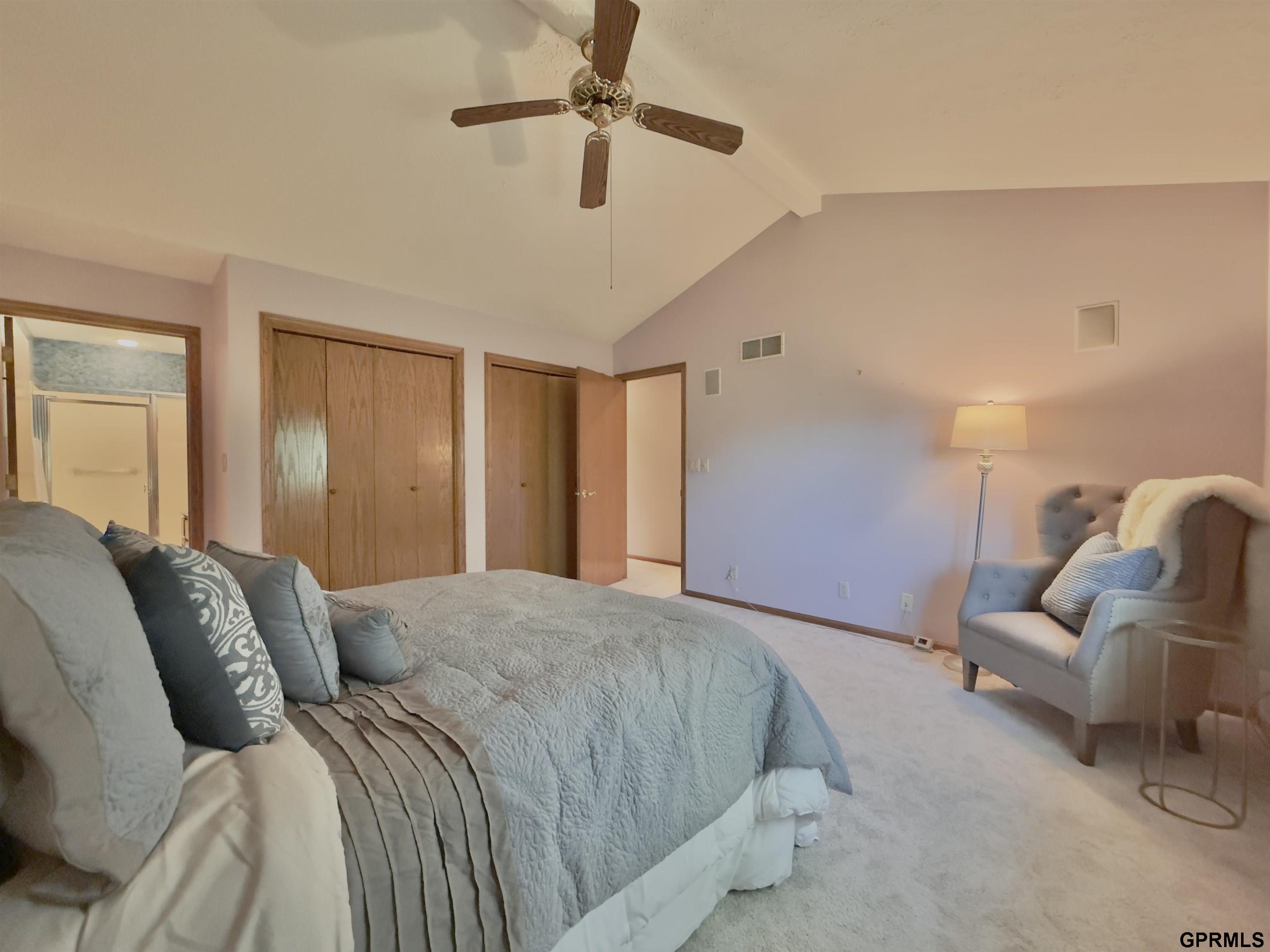 Furnished bedroom with a blue-gray quilted bed, tufted armchair, ceiling fan, and wooden closet doors.