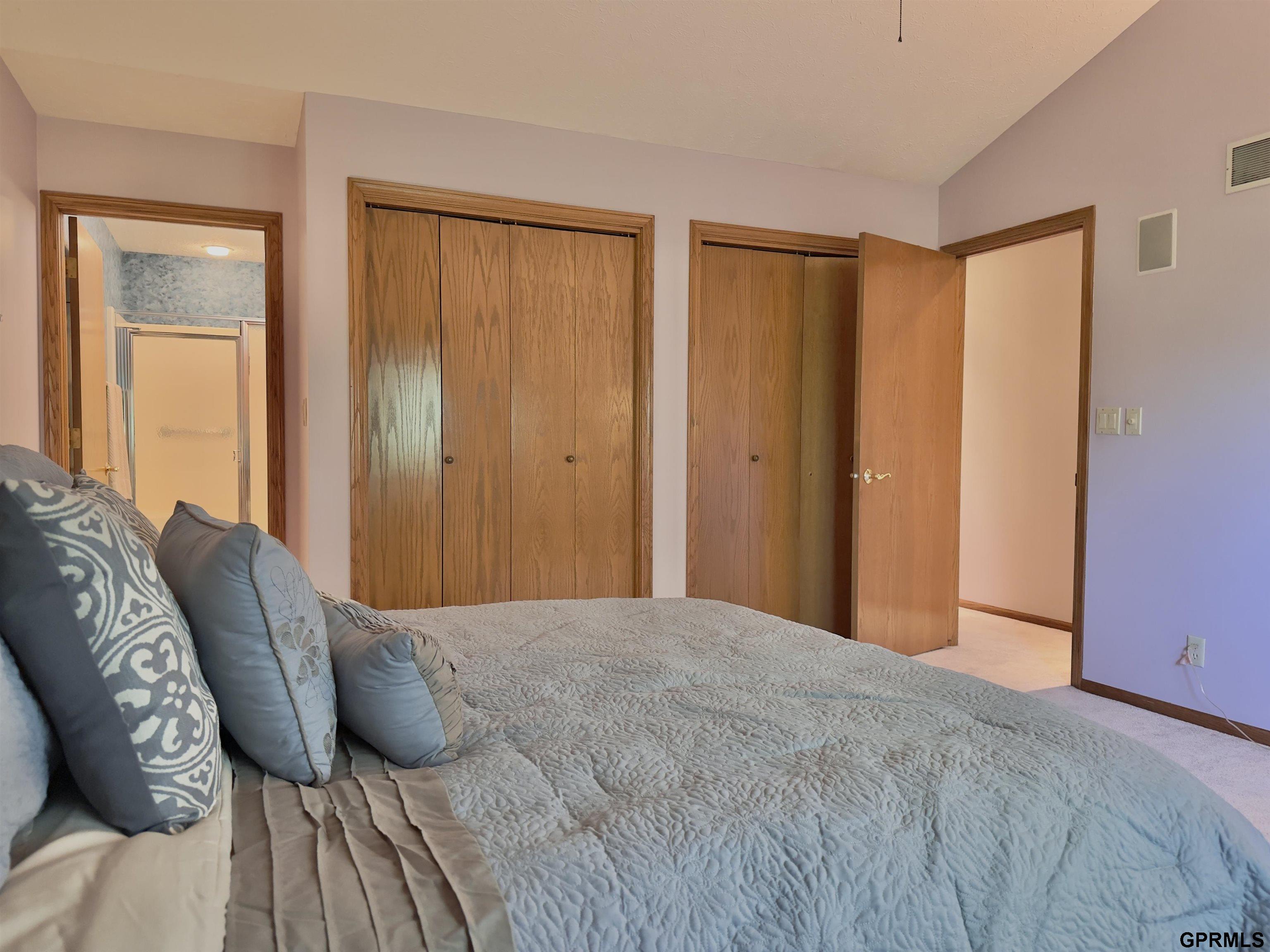 Bedroom with a large gray-quilted bed, wood closet doors, and an adjoining bathroom visible through the doorway.
