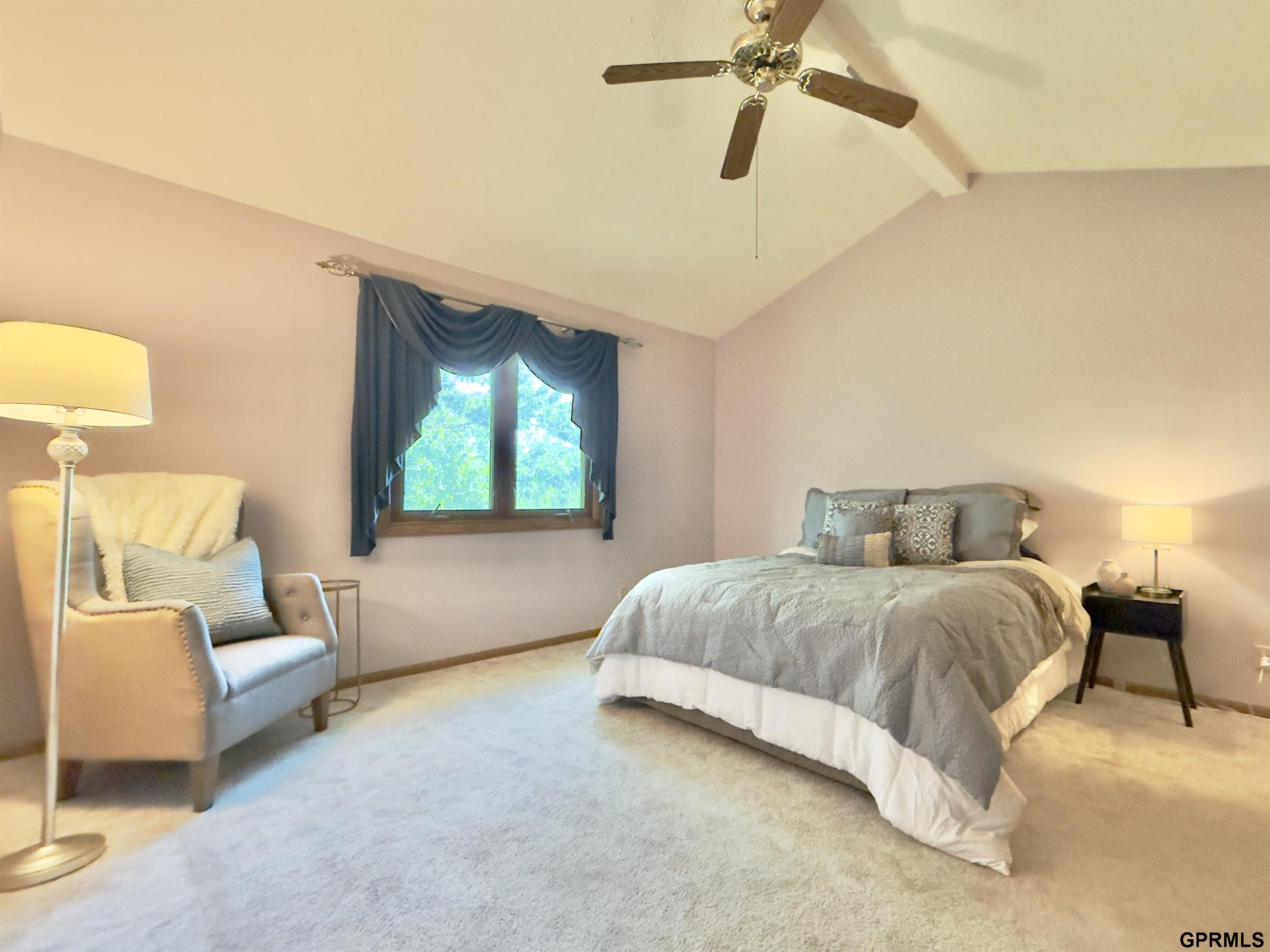 Furnished bedroom with a large gray-covered bed, an armchair, and a vaulted ceiling with a ceiling fan.
