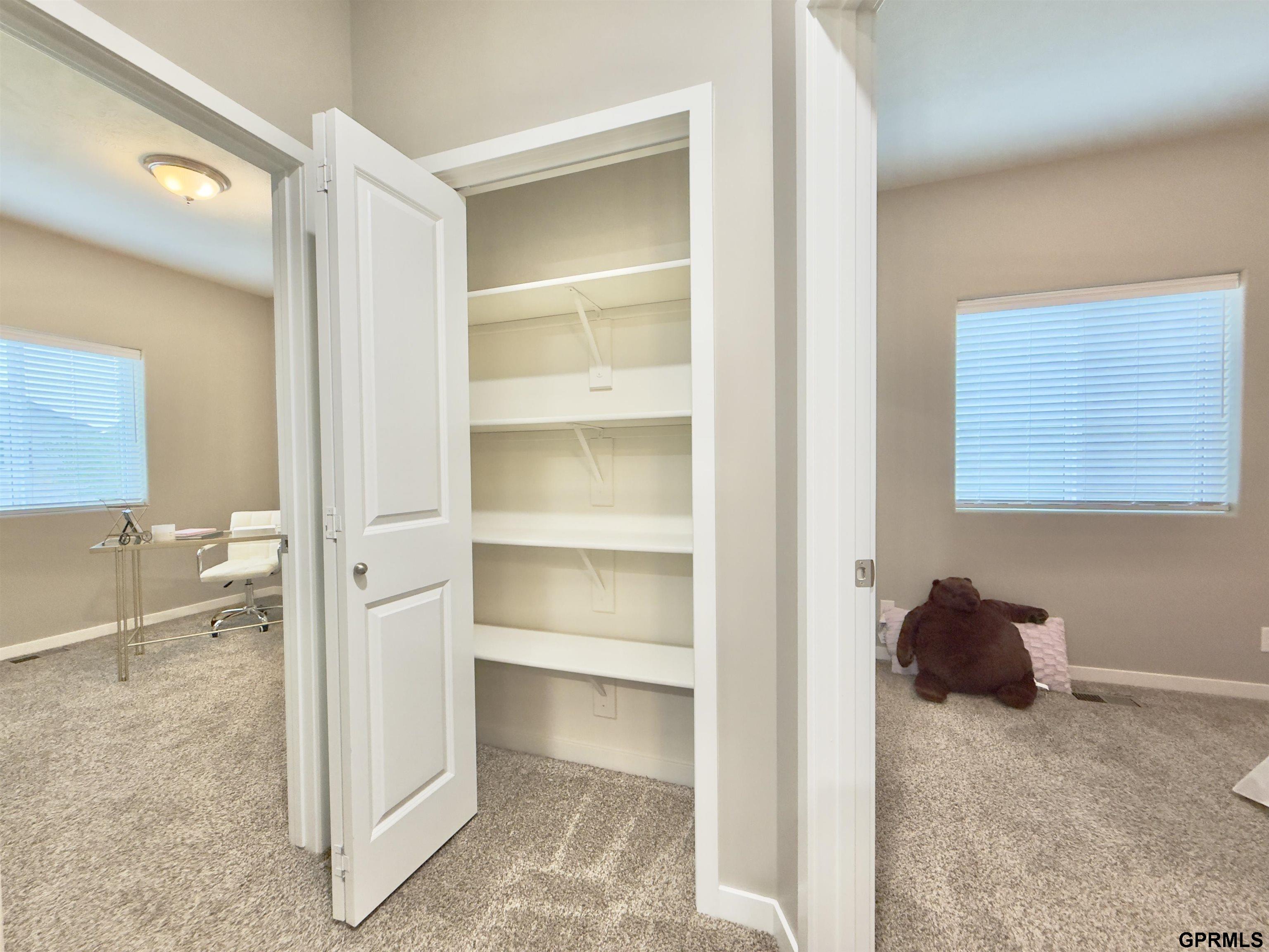 Open closet with multiple white shelves between two carpeted rooms.