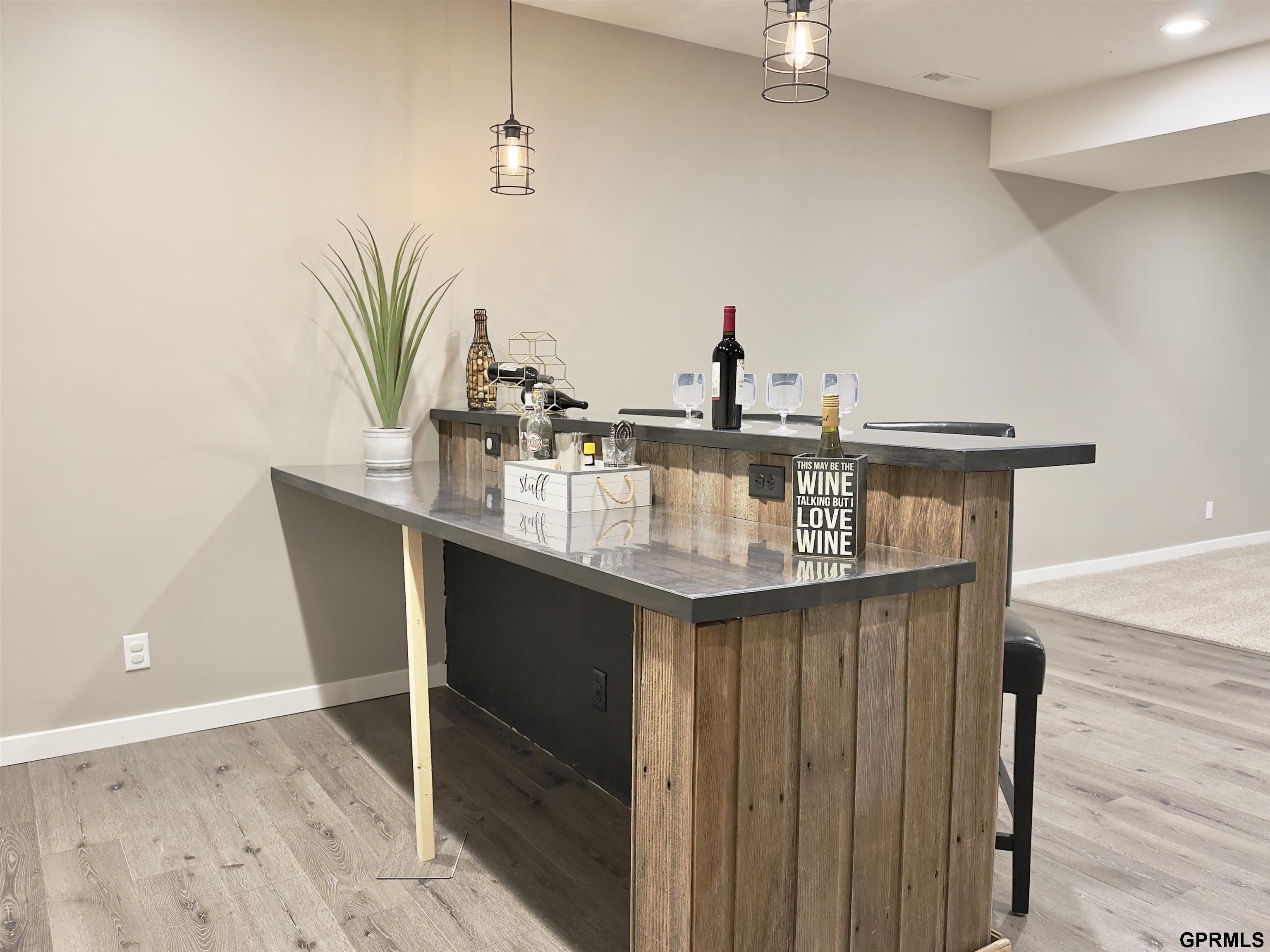 Basement bar with rustic wood-front counter, wine bottles and glasses, and two hanging pendant lights.