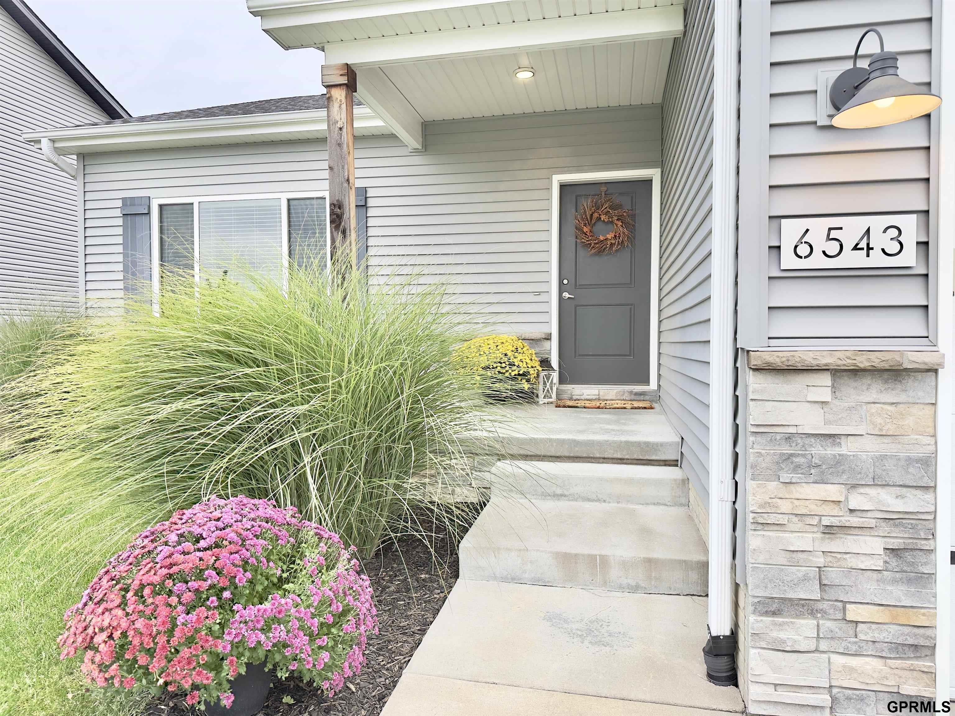 Gray-sided home entrance with stone veneer, concrete steps, and colorful flower beds; house number 6543.
