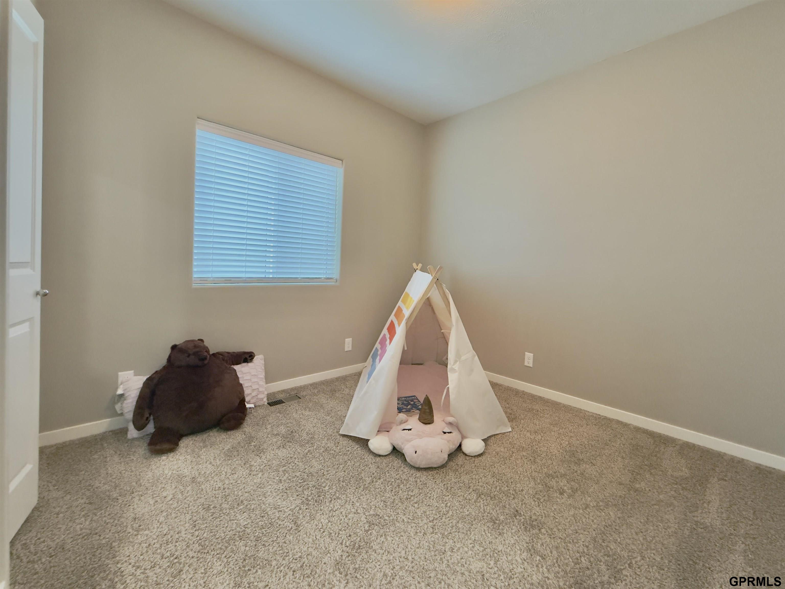 Carpeted bedroom playroom with a small teepee tent and stuffed animals beneath a window with closed blinds.
