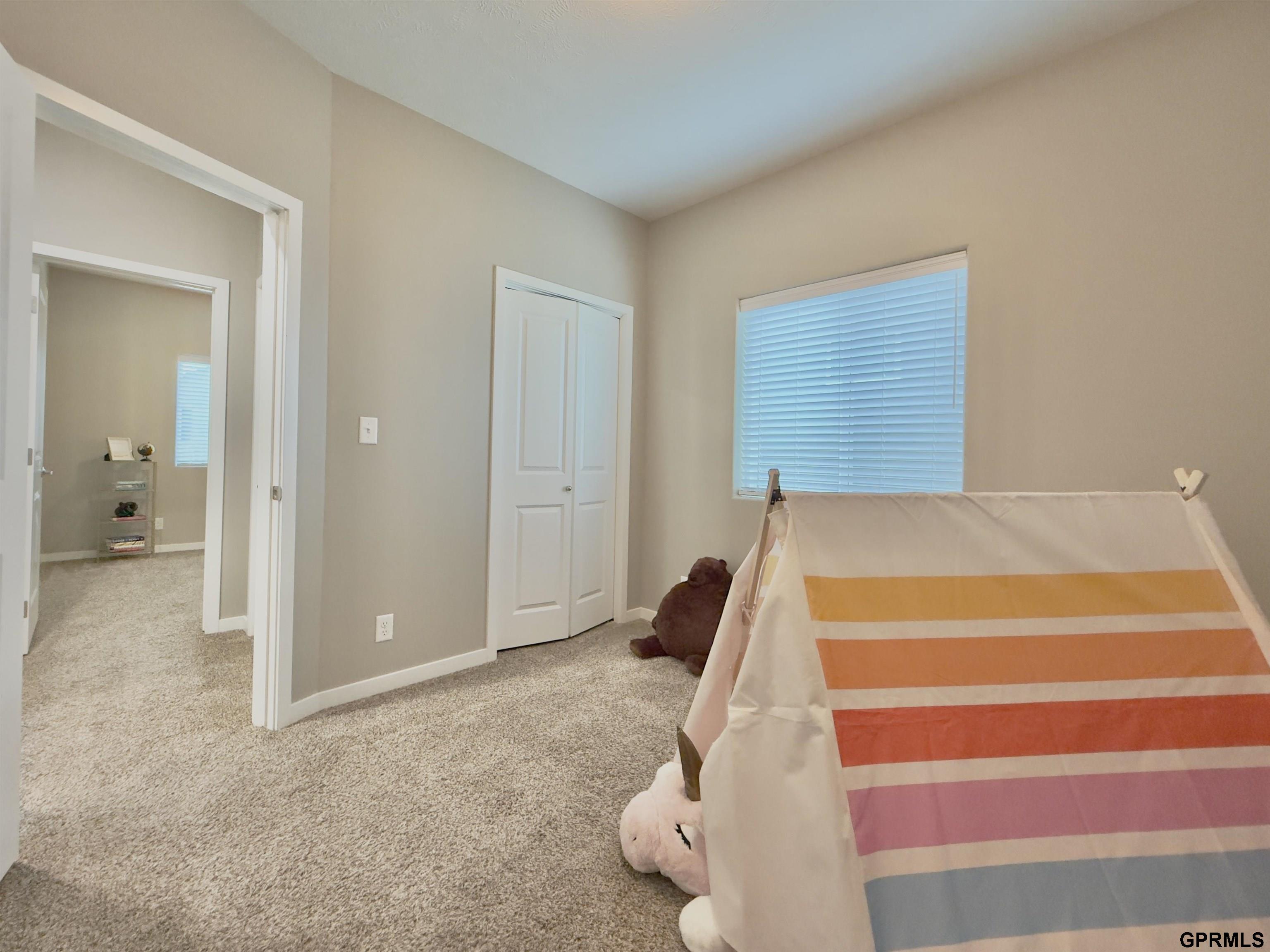 Carpeted bedroom with a colorful striped play tent, stuffed animals, and a window with blinds.