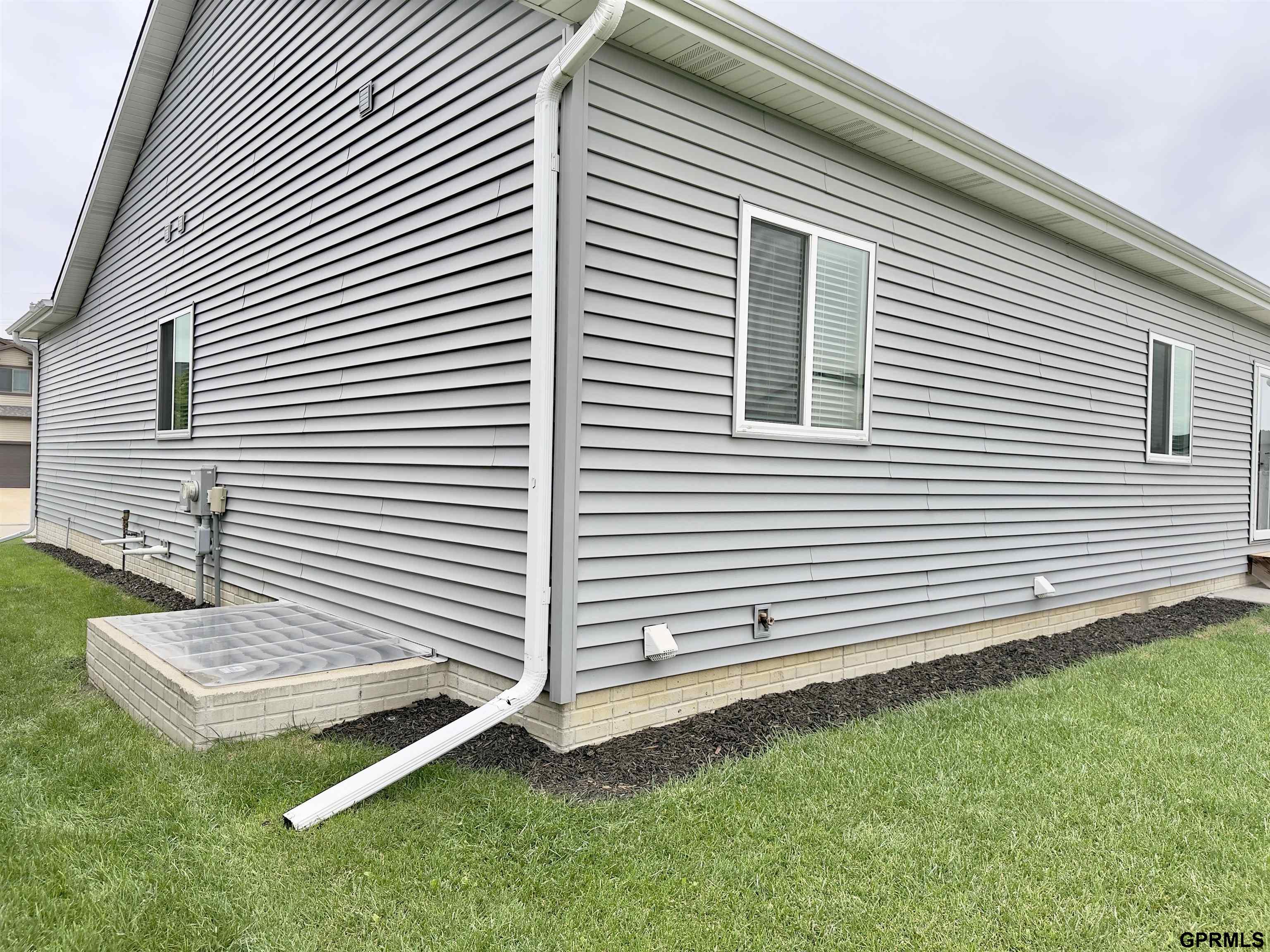 House side with gray siding, brick foundation, and a detached downspout extension across the lawn.