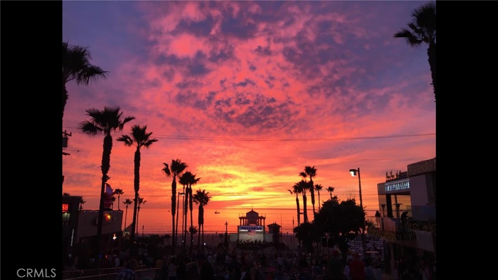 Sunset over the ocean, framed by palm trees and beachside buildings.