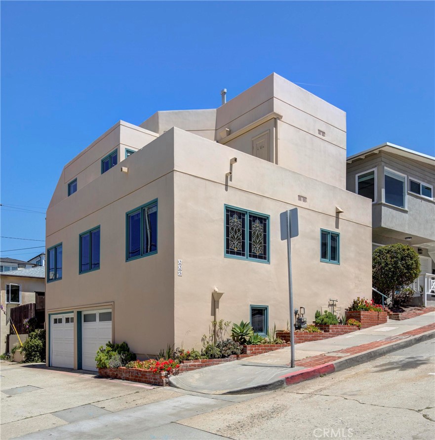 Modern multi-story stucco home with teal-trimmed windows, two garage doors, and brick-edged flower beds.