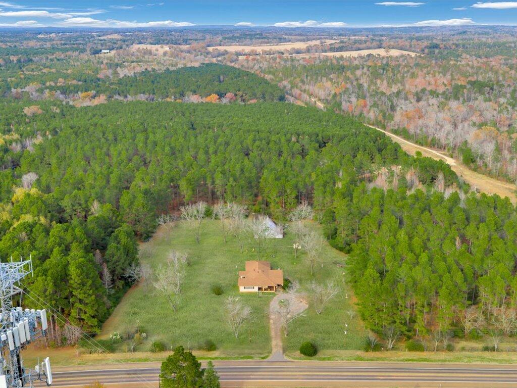 Aerial view of a house in a wooded clearing beside a highway, with a long driveway and surrounding acreage.