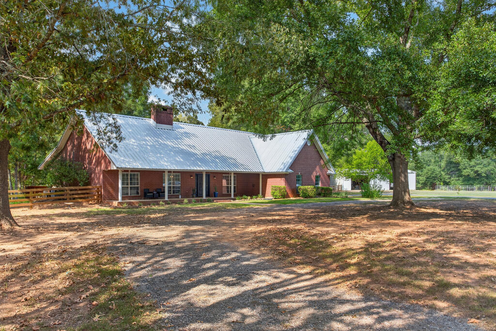 Single-story brick house with a light metal roof beneath large shade trees, beside a gravel driveway.