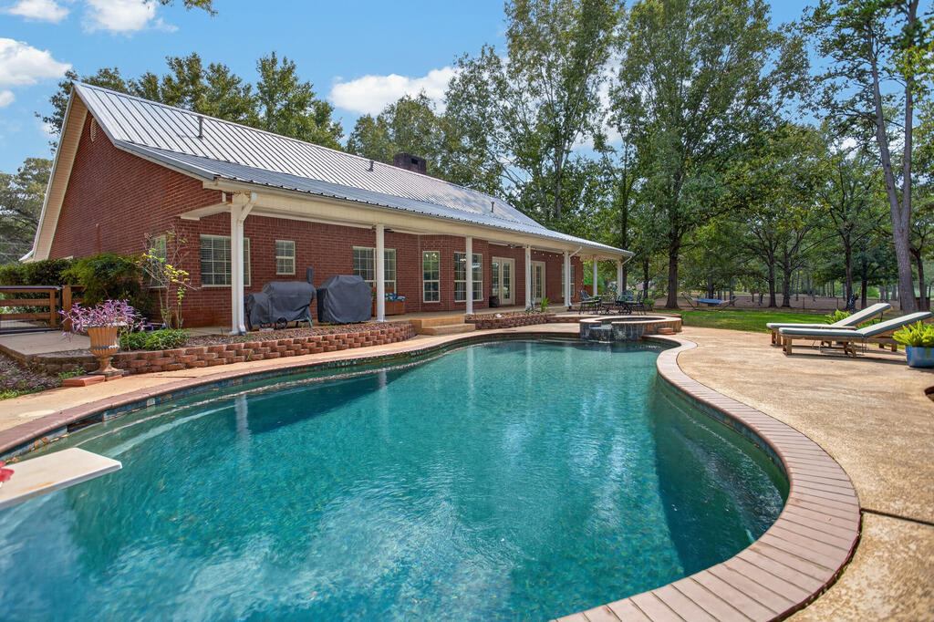 Curved backyard swimming pool beside a brick home with a metal roof and patio seating.