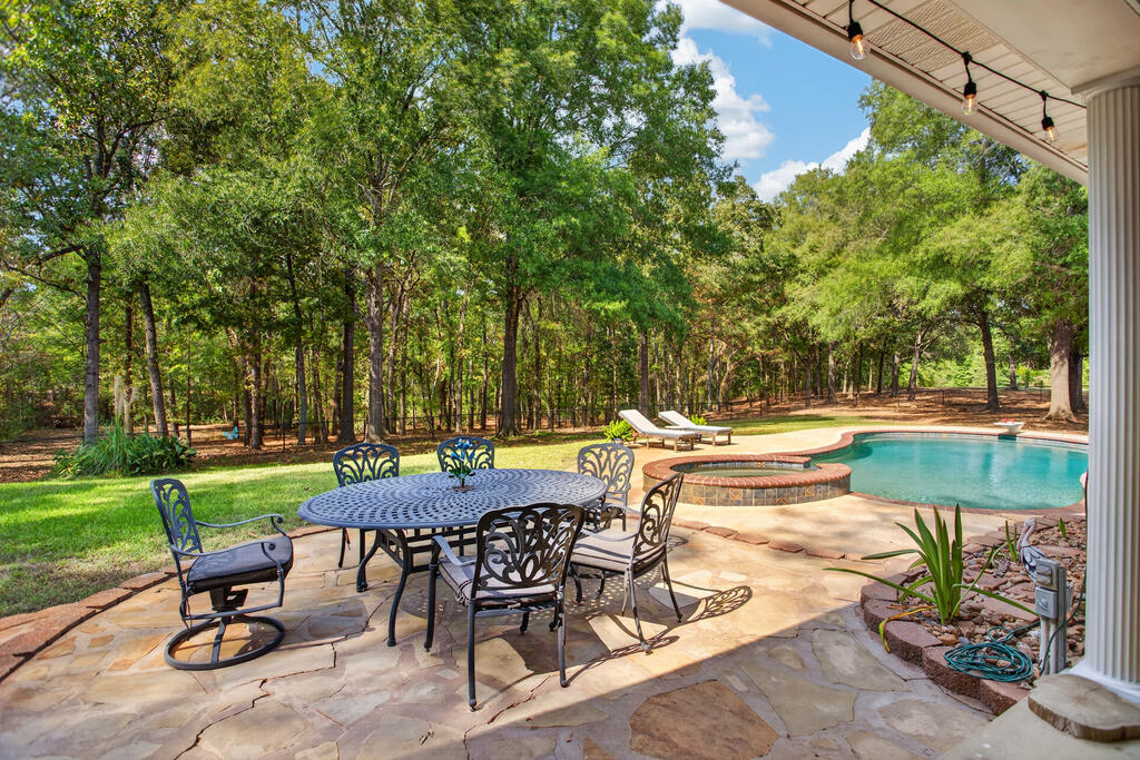 Swimming pool beside a stone patio with outdoor dining furniture and a wooded yard.
