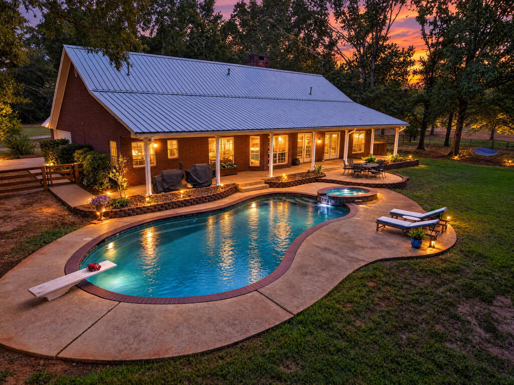Brick ranch home with a metal roof beside a curved pool and attached spa at dusk.