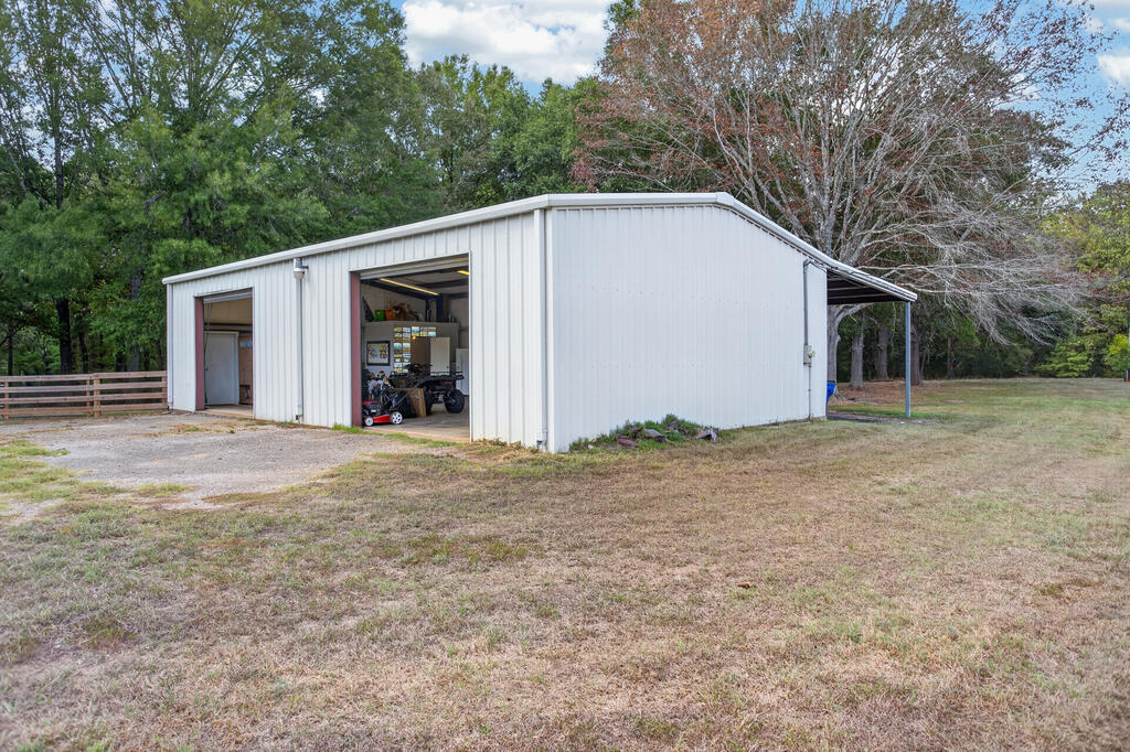 White metal workshop with open garage bays and a covered side awning in a grassy, wooded yard.