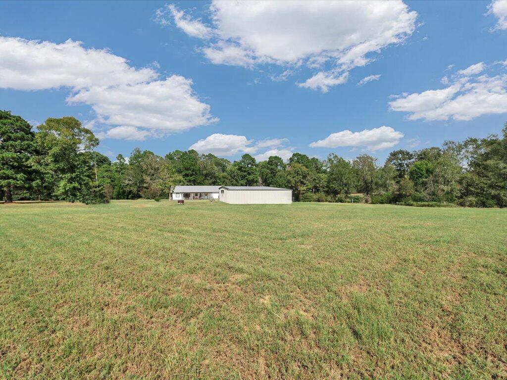 Small single-story home and white metal outbuilding beyond a wide grassy field and wooded tree line.