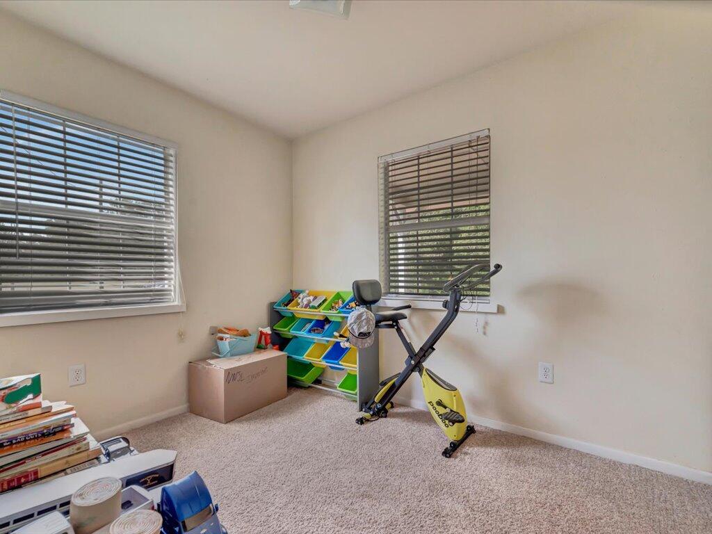 Carpeted recreation room with an exercise bike, colorful toy organizer, and two windows with blinds.