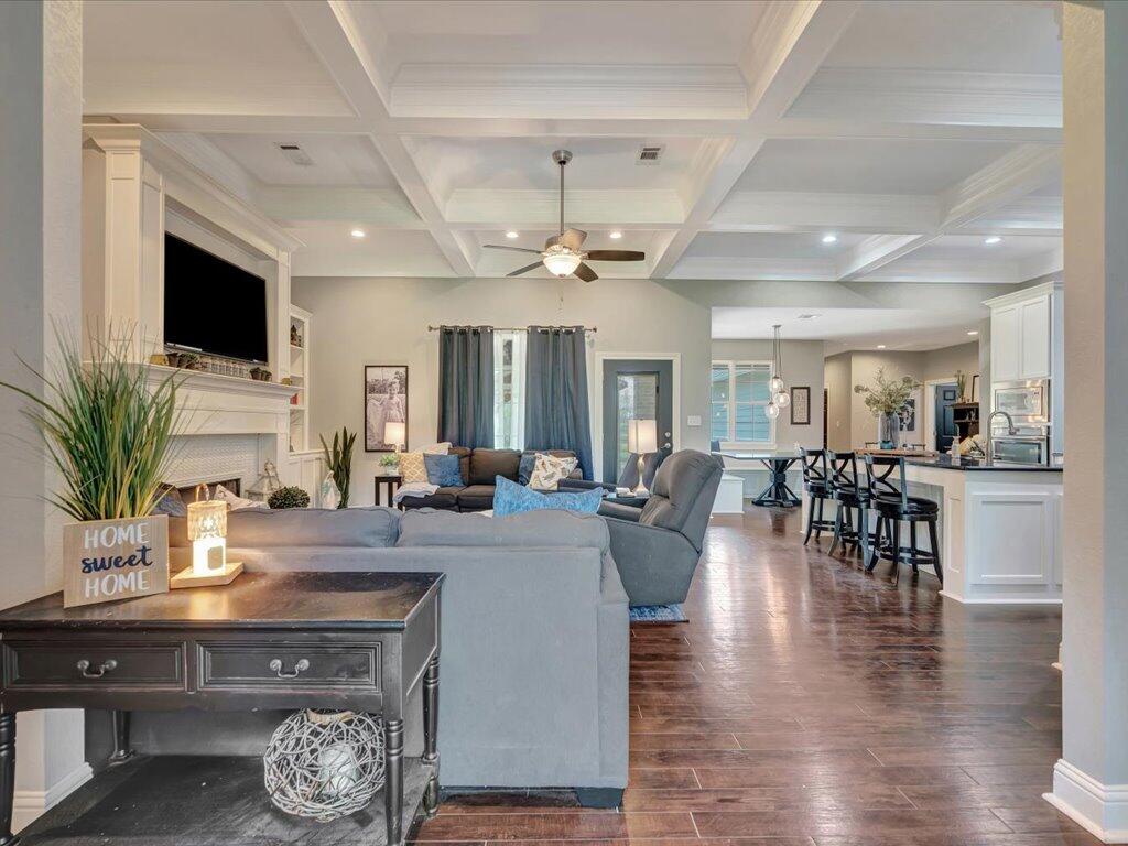 Open living room with coffered ceiling, fireplace, dark wood floors, and adjoining kitchen.