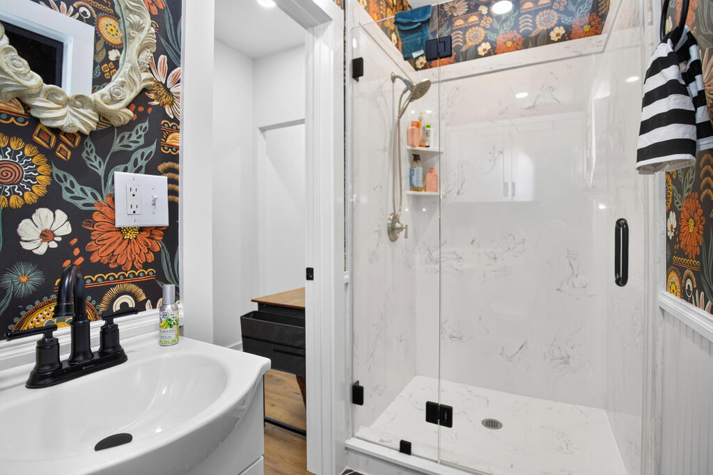 Bathroom with a glass-enclosed marble-look shower, white vanity, and bold floral wallpaper.