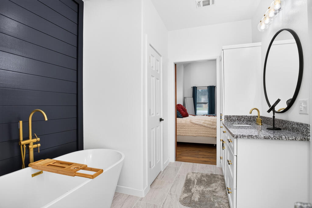 Bathroom with freestanding tub, dark shiplap wall, gold fixtures, and vanity beside a bedroom doorway.