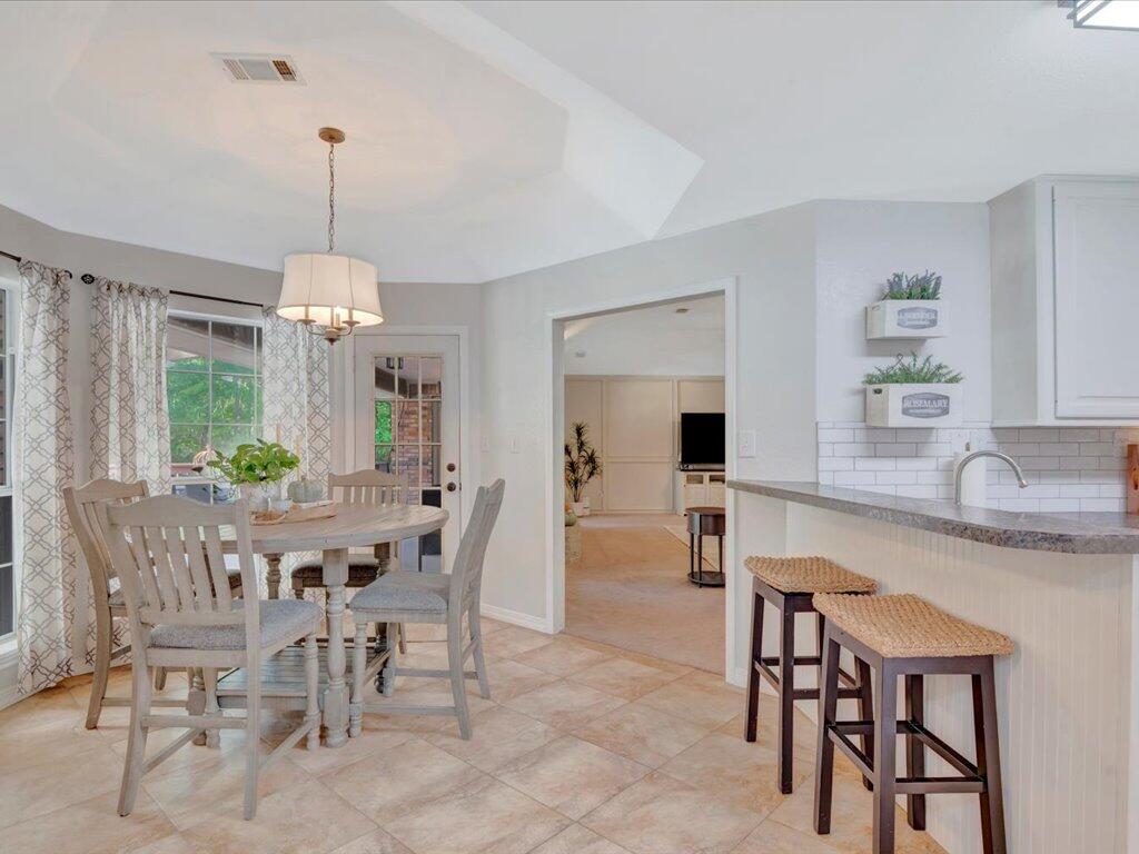 Dining area with a round table beside large windows, opening to a kitchen with a breakfast bar.