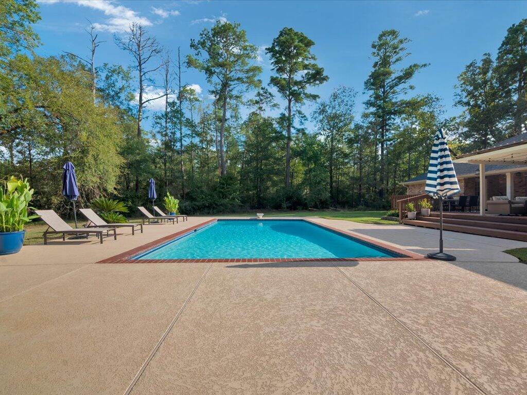 Backyard swimming pool with a wide concrete deck, lounge chairs, and wooded surroundings.