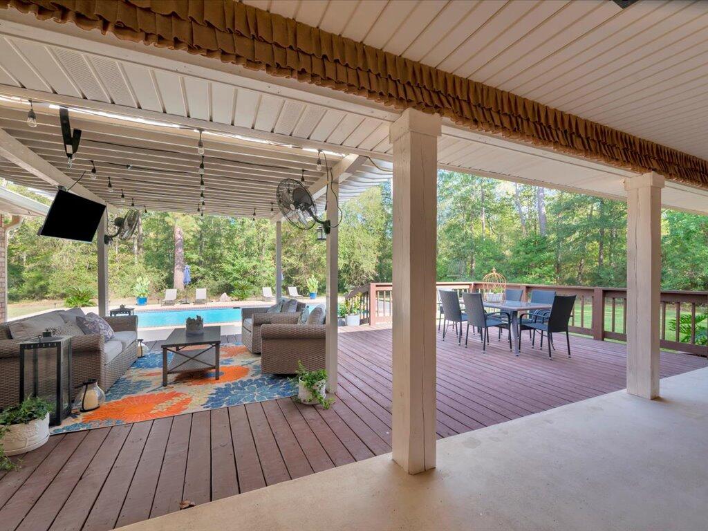 Covered deck with lounge seating and dining area beside a swimming pool and wooded yard.