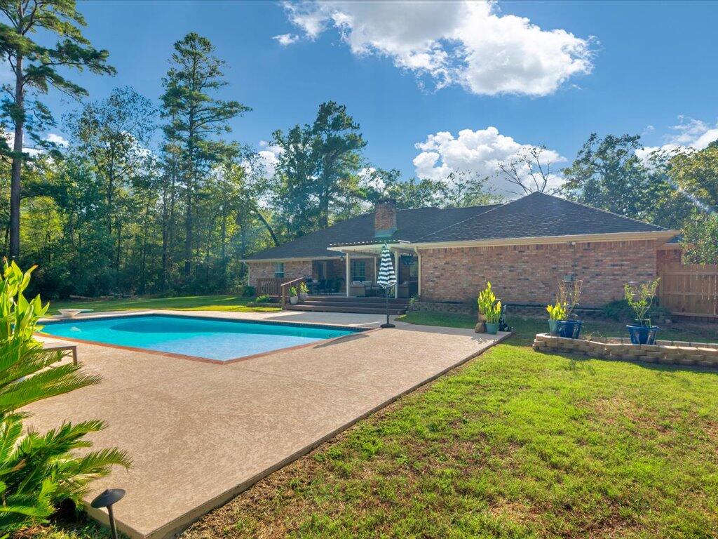 Brick home behind a swimming pool and wide paved deck, with a covered patio and wooded yard.