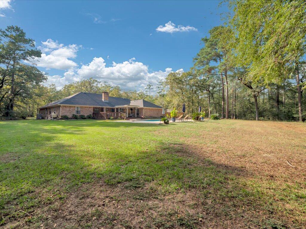 Brick home beyond a broad lawn, with a backyard pool, patio seating, and wooded surroundings.