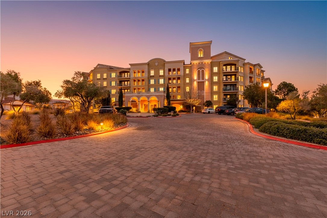 Multi-story building with balconies and illuminated windows at dusk, viewed from its landscaped entrance drive.