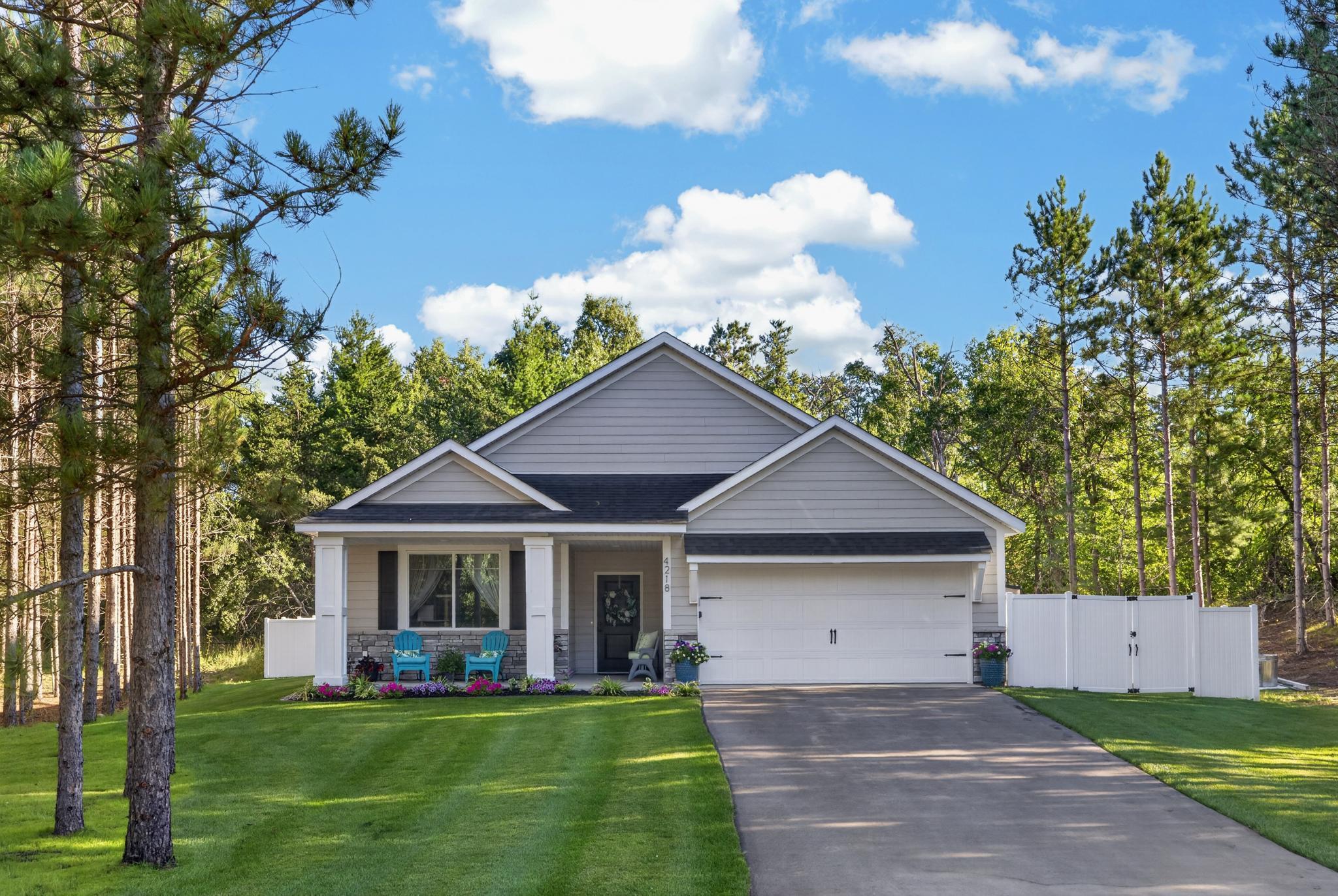 Gray home with a two-car garage, covered porch, and white fence among tall trees.