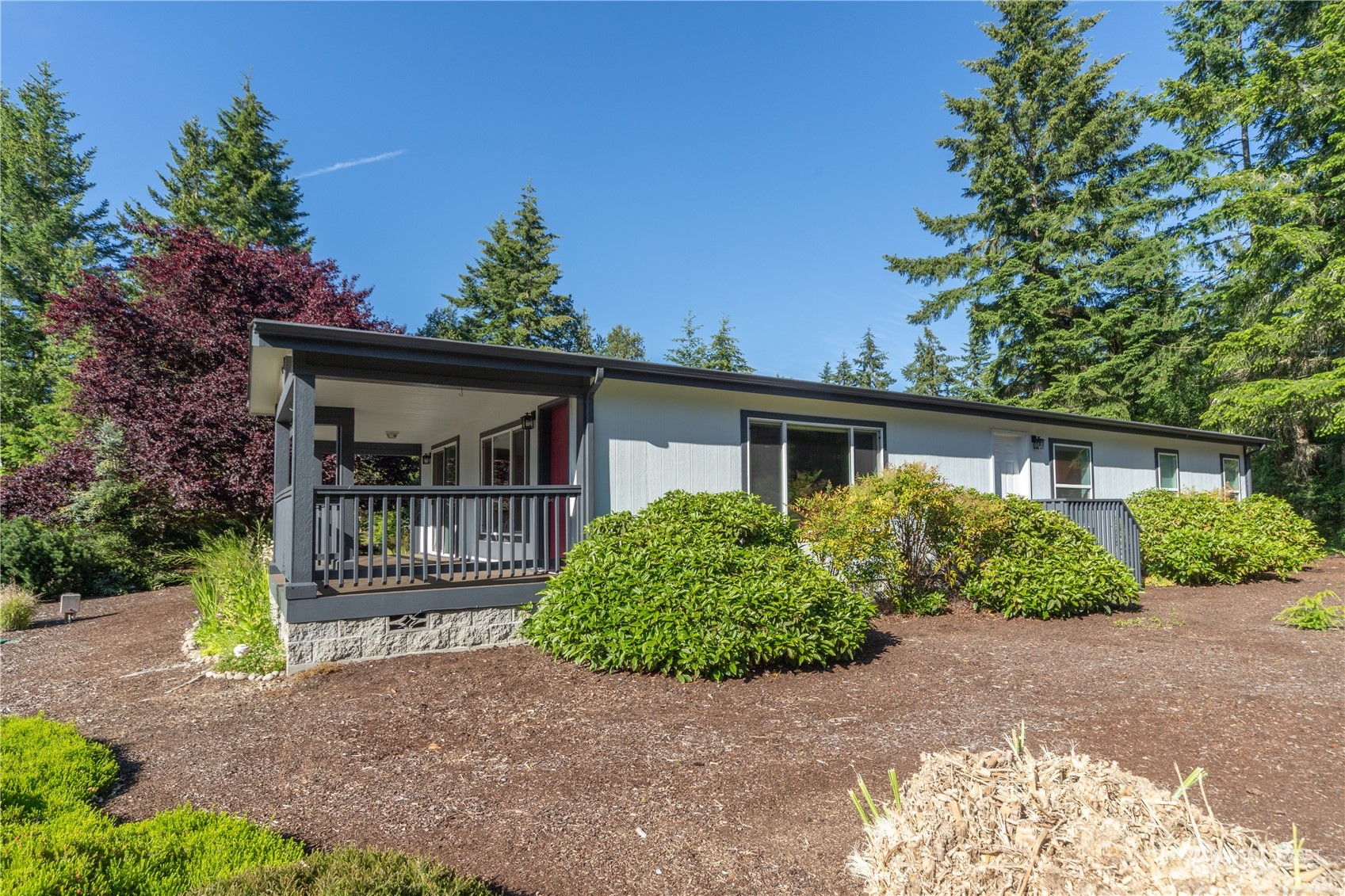 Single-story gray home with a covered porch, dark railings, and landscaped shrubs among evergreen trees.