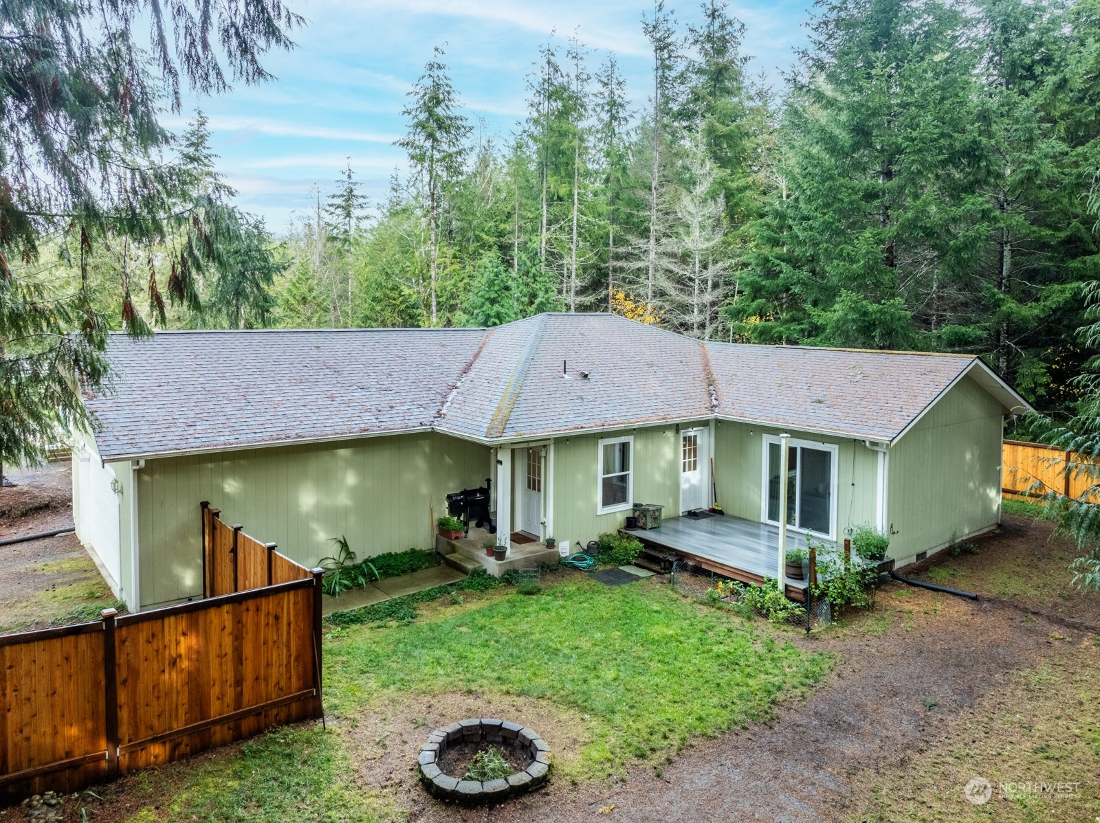 Sage-green single-story home with a fenced yard and circular stone fire pit, surrounded by evergreen woods.