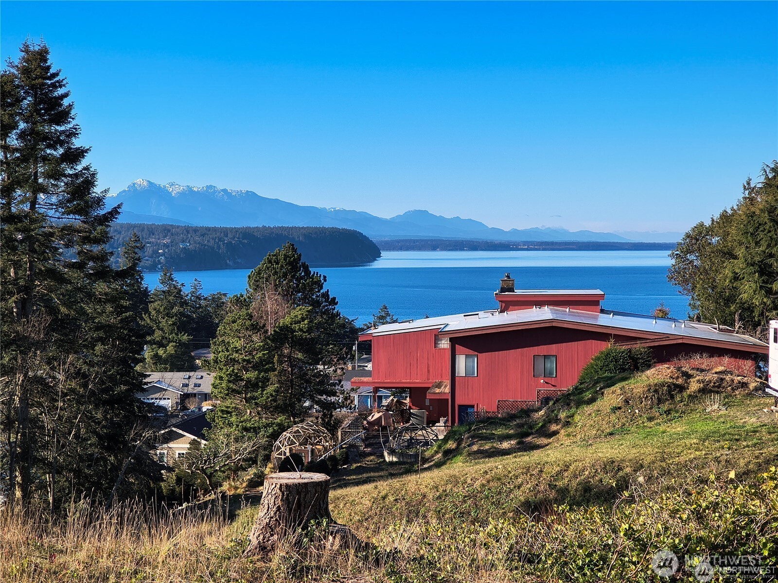 Red hillside home overlooking blue water and distant snow-dusted mountains.