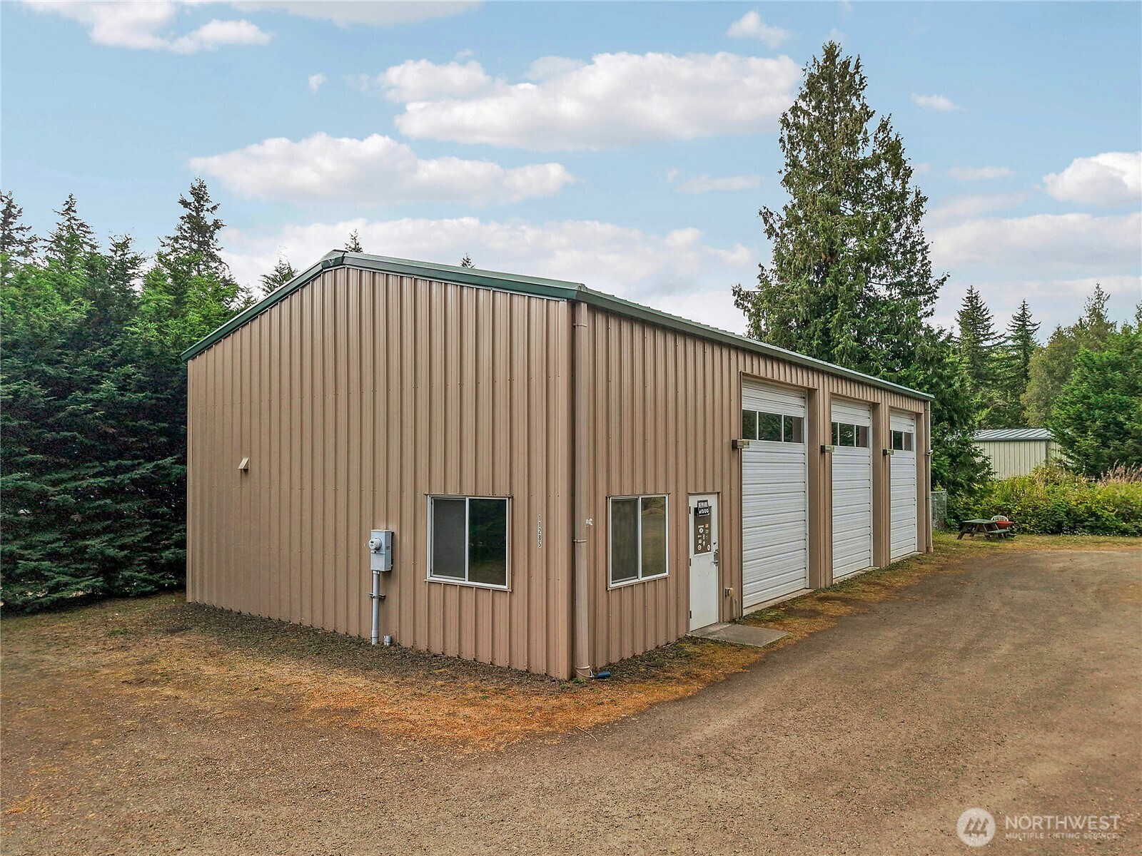 Brown metal workshop with three roll-up garage doors beside a gravel drive and evergreen trees.
